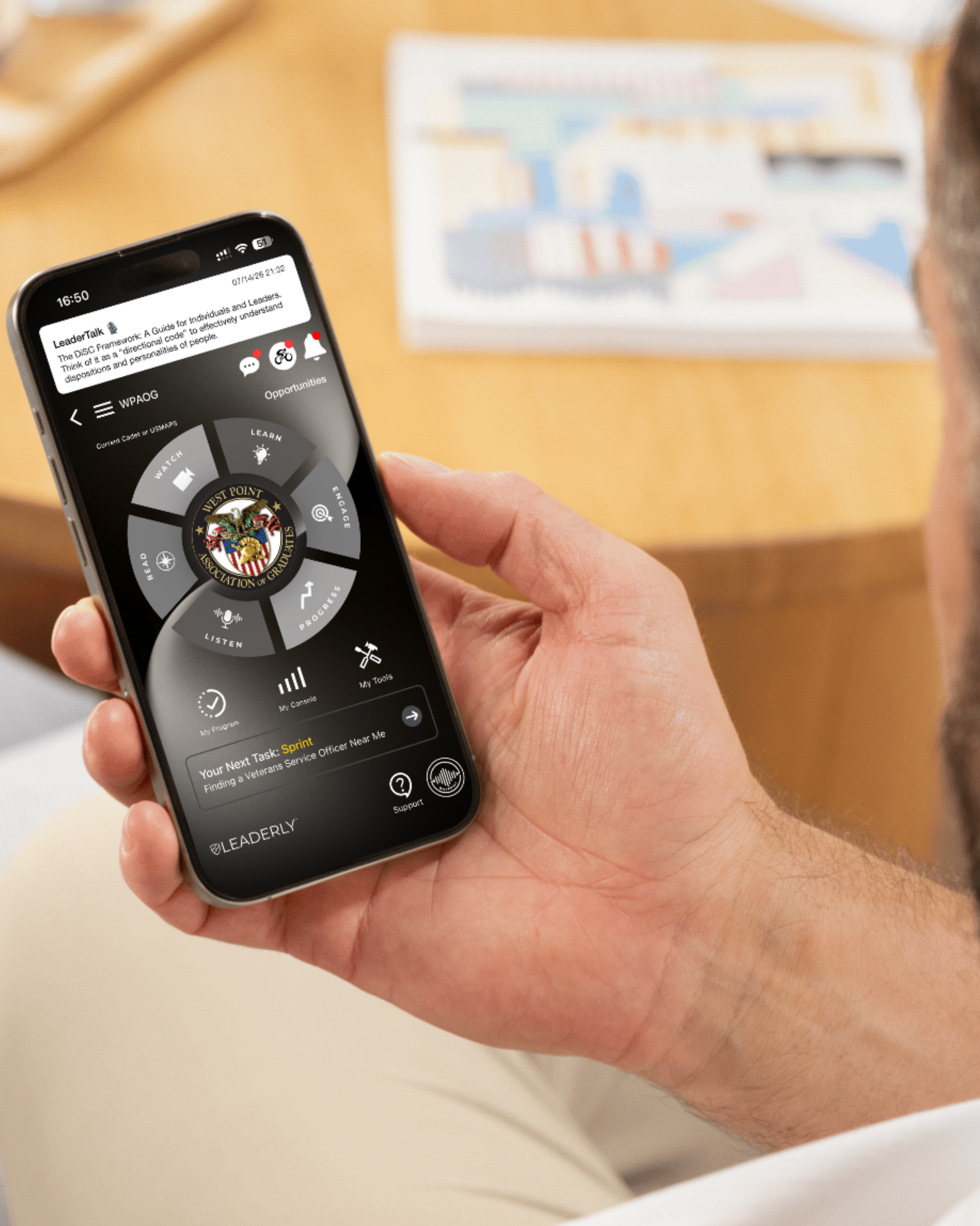 Close-up of a person's hand holding a smartphone, displaying a circular app interface with a veterans organization emblem in the center. The background includes a blurred diagram or map.