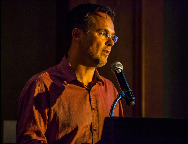 A man wearing glasses and a red button-up shirt stands at a microphone, appearing to give a speech or presentation in a dimly lit indoor setting.