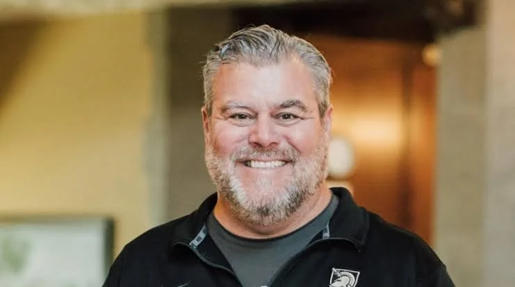 A smiling man with gray hair and a beard, wearing a black jacket indoors.