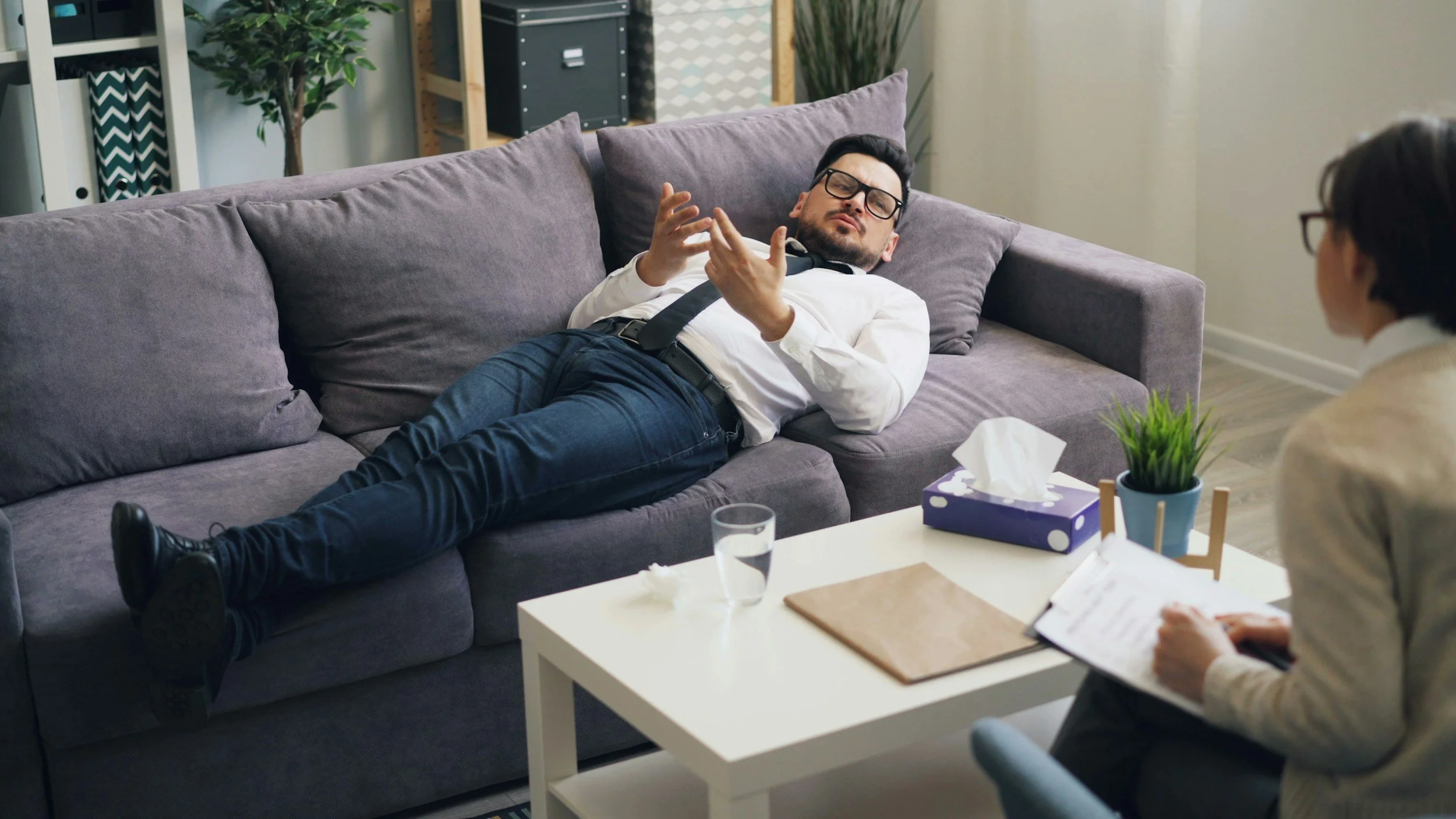 A man with dark hair, glasses, and a beard lies on a gray couch, gesturing with his hands while talking to a woman sitting across from him. The man is wearing a white shirt and blue pants, and has a black tie loosened around his neck. The woman has short dark hair and glasses, wearing a light-colored blazer, and is taking notes on a clipboard. A white coffee table in front of them holds a tissue box, a glass of water, and a small plant in a blue pot.