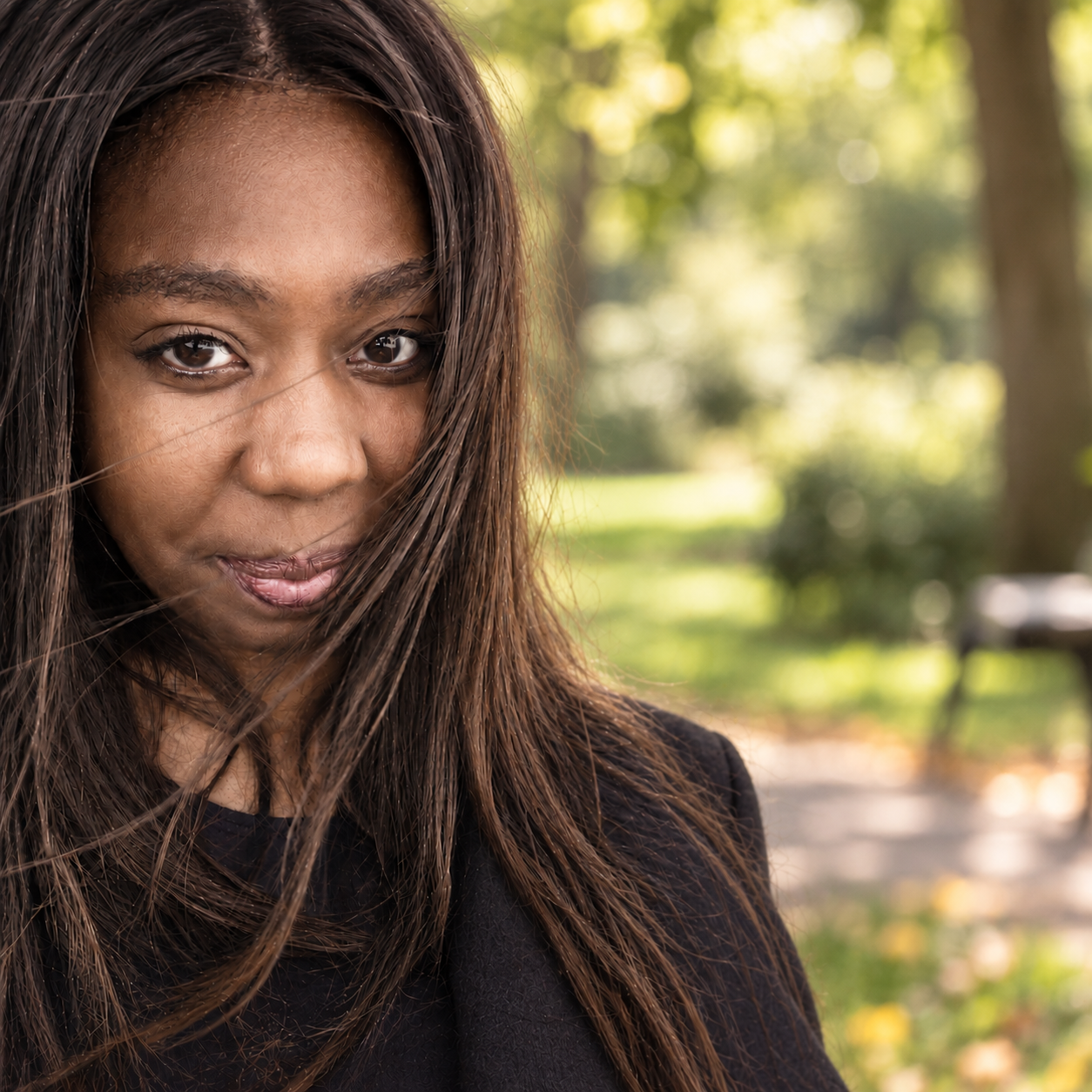Close-up of a woman with long brown hair, looking into the camera with a subtle smile, outdoors in a park with sunlight filtering through trees.