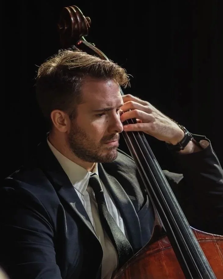 A man with brown hair and a beard playing a double bass, wearing a black suit, white shirt, and black tie, with a serious expression, against a dark background.