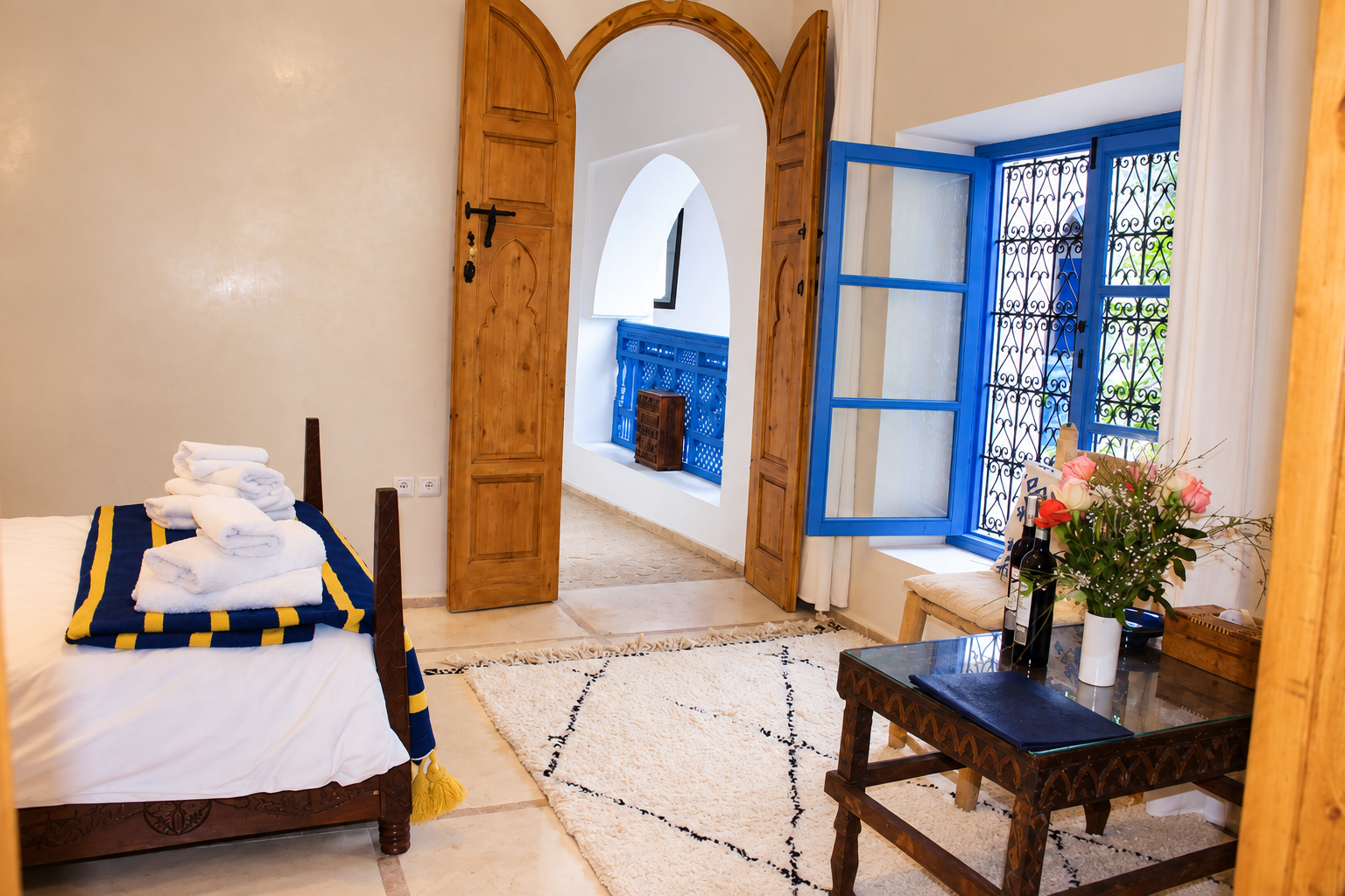 A cozy hotel room with a bed topped with folded towels, a wooden table with flower vase, a blue window with intricate metal grill, and a doorway leading to a balcony with a decorative blue railing.