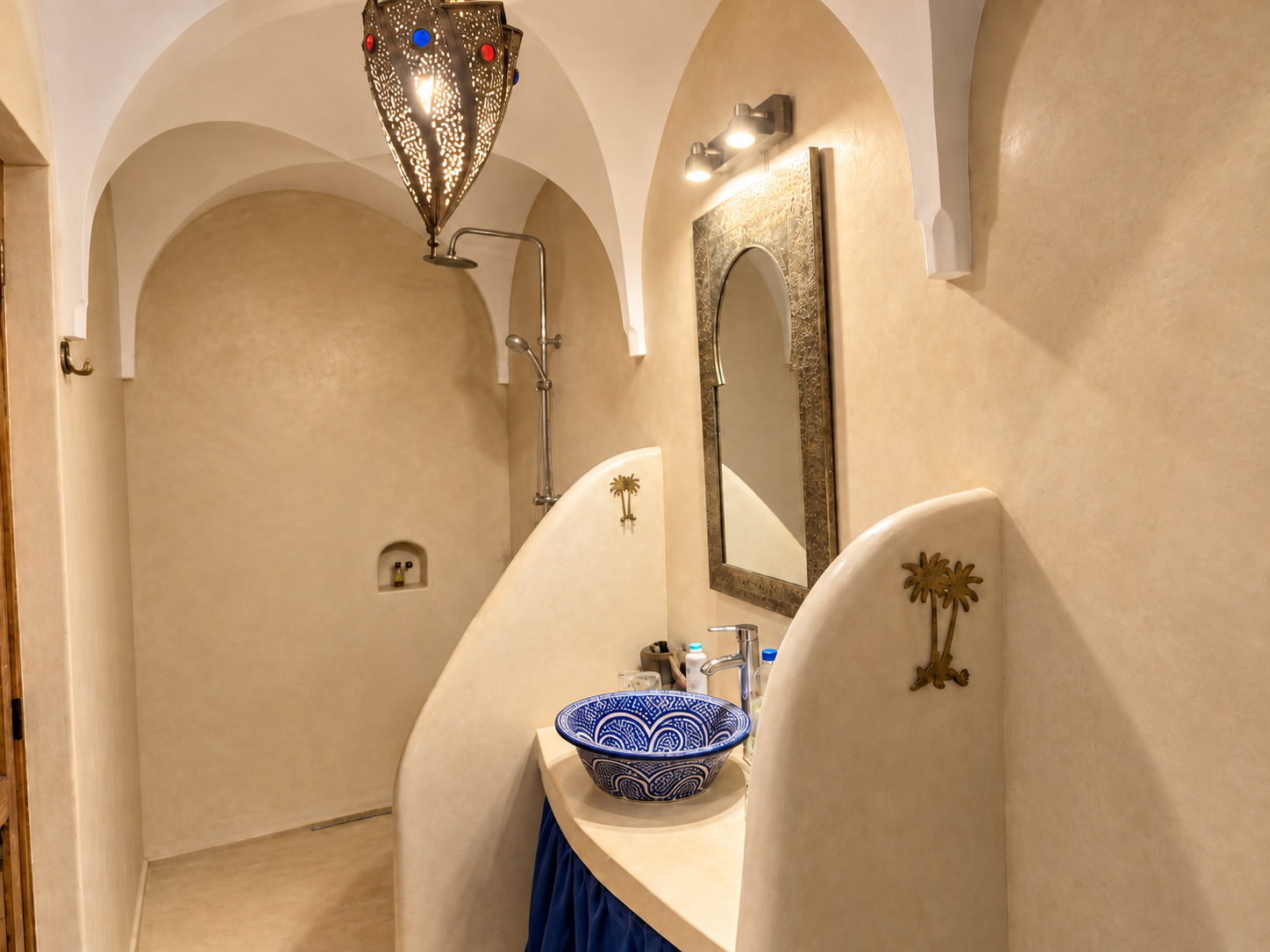 A bathroom featuring a blue decorative sink, a large ornate mirror, and Moroccan-style hanging lamp, with curved partition walls, beige textured walls, and a walk-in shower with a rain showerhead.