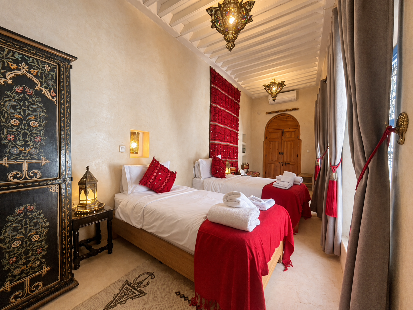 A bedroom with two twin beds featuring red bedspreads and pillows, a patterned black wardrobe, beige walls, and a door at the end of the room. The room is decorated with Moroccan-style lamps and curtains.