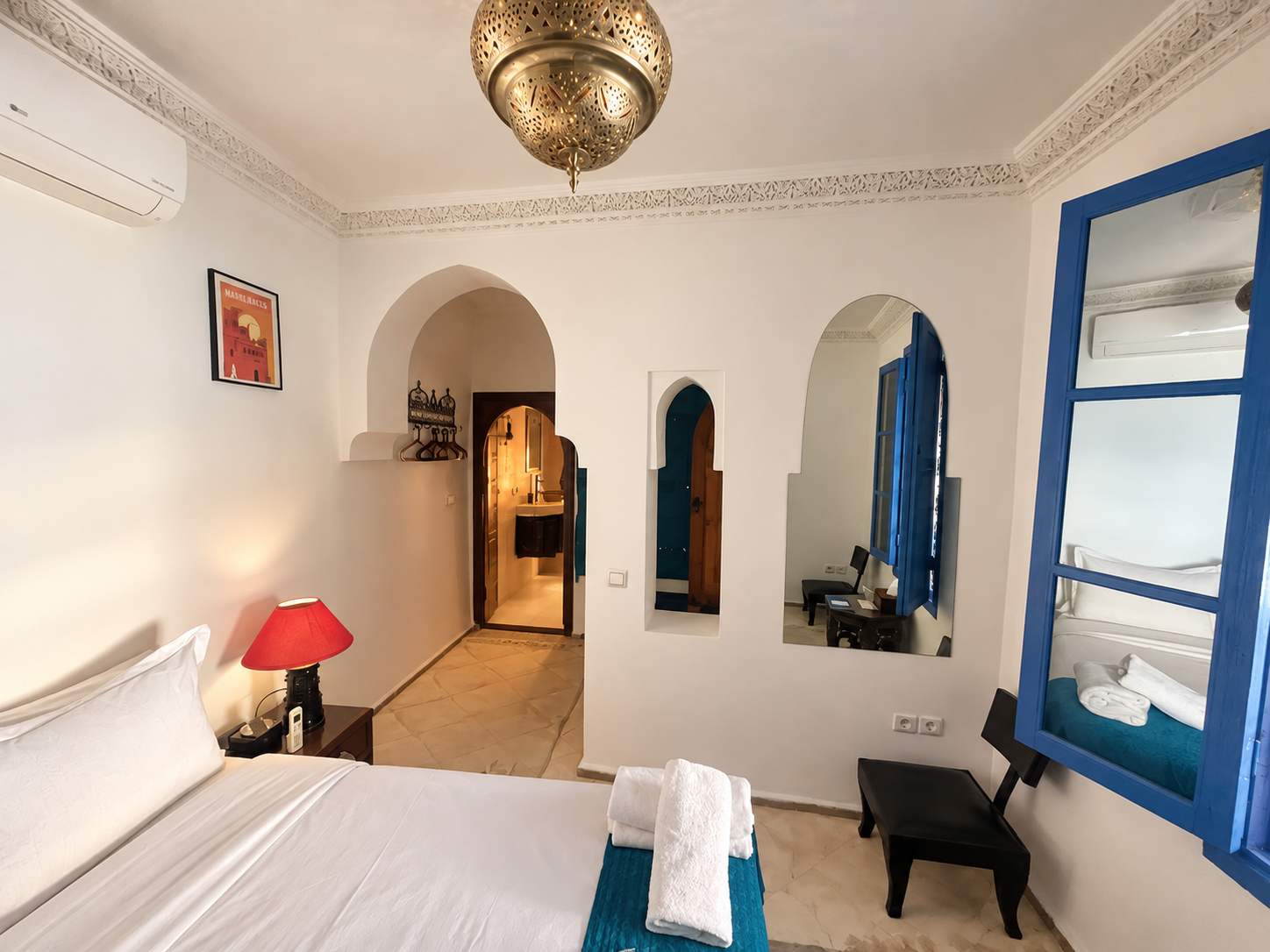 A cozy hotel room with white walls and blue accents. It features a bed with white linens and a rolled towel on top. A red bedside lamp, a black chair, and mirrors on the wall are visible. The room has an archway leading to a small bathroom area, with a window with blue shutters next to the bed.