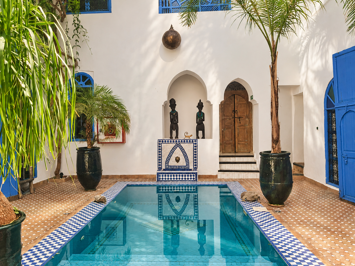 A courtyard with a swimming pool in front, surrounded by terra cotta tile flooring, large potted plants, and white walls. There are three black wooden statues with turquoise necklaces on a ledge, and a wooden door to the right. Blue shutters and window frames are visible, with sunlight casting shadows.