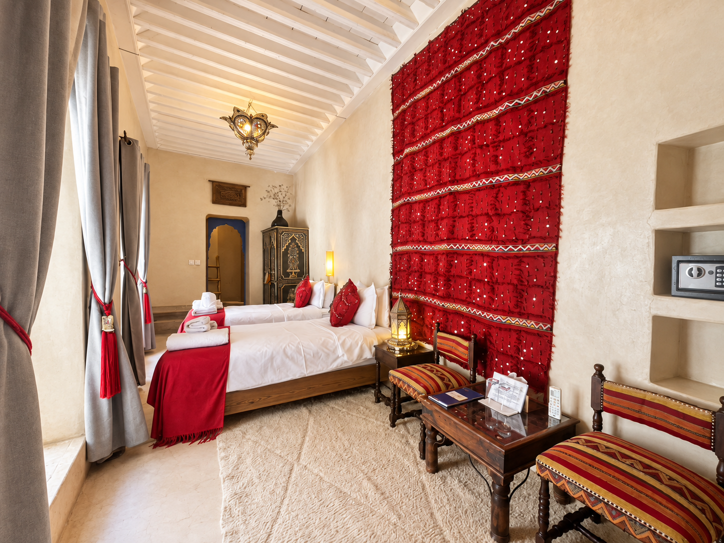 A hotel room with two beds decorated in red and white, wooden furniture, patterned chairs, a large red wall hanging, and Moroccan-style lighting.