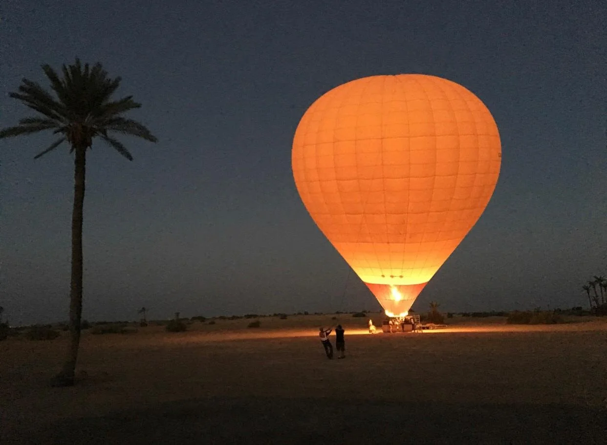 A hot air balloon glowing orange at night on a flat landscape with a single palm tree in the foreground.