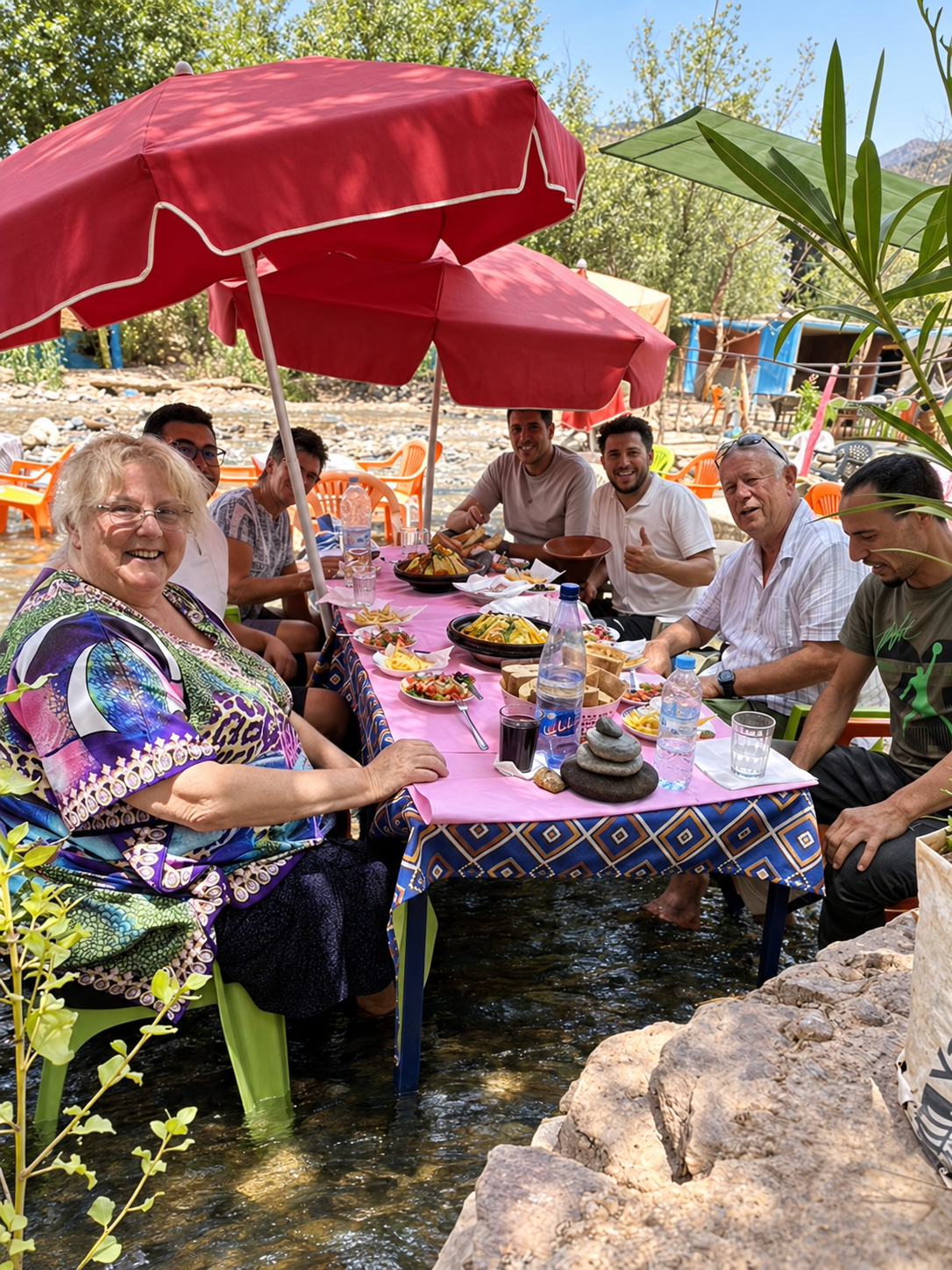 A group of people sitting at a table outdoors, enjoying a meal under large red umbrellas with a river beneath the table and surrounding trees.