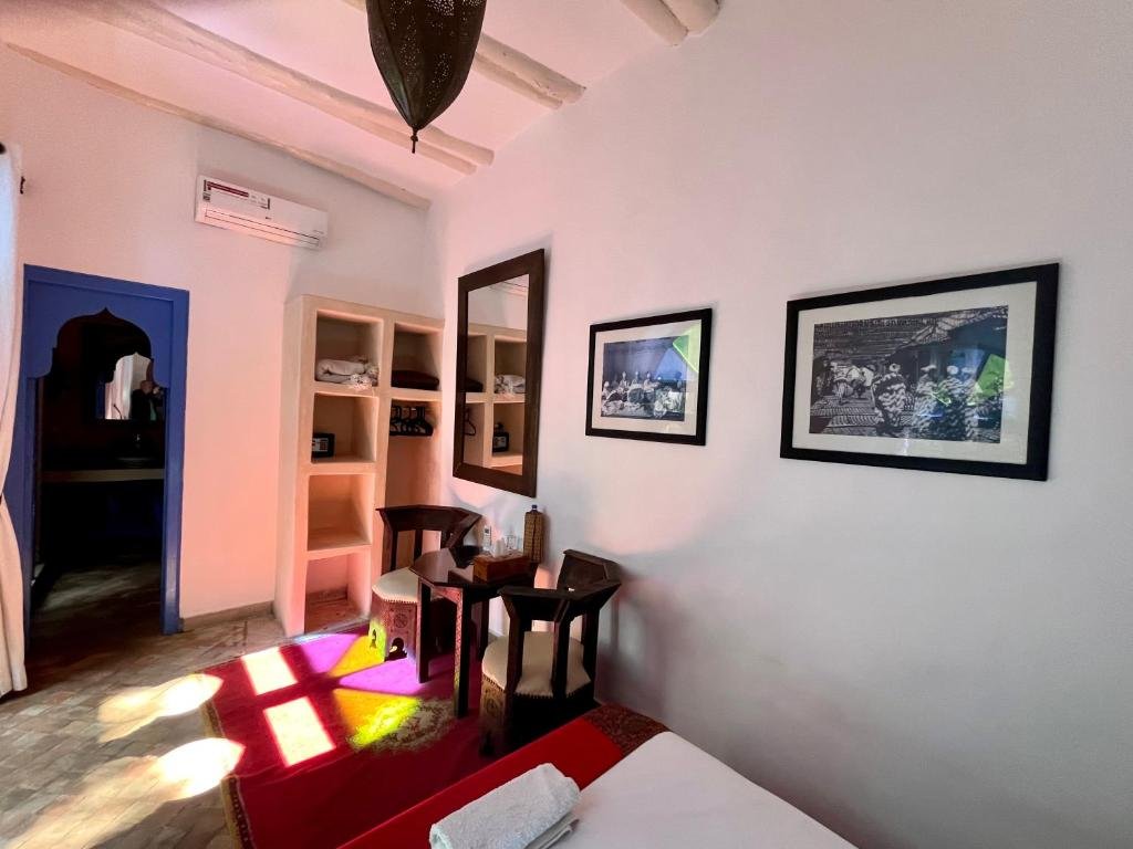 A hotel room with a white wall decorated with two black-framed photos, a wooden mirror, a small table with two chairs, bed with folded towels, a built-in shelf with towels and small items, a blue door frame, and a black hanging light fixture. Sunlight creates a pink and yellow pattern on the floor.