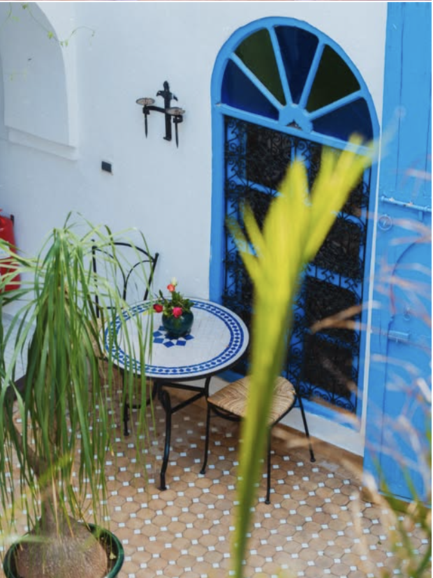 Outdoor patio with a small round table decorated with flowers, two chairs, a potted plant, and a blue shuttered window on a white wall.