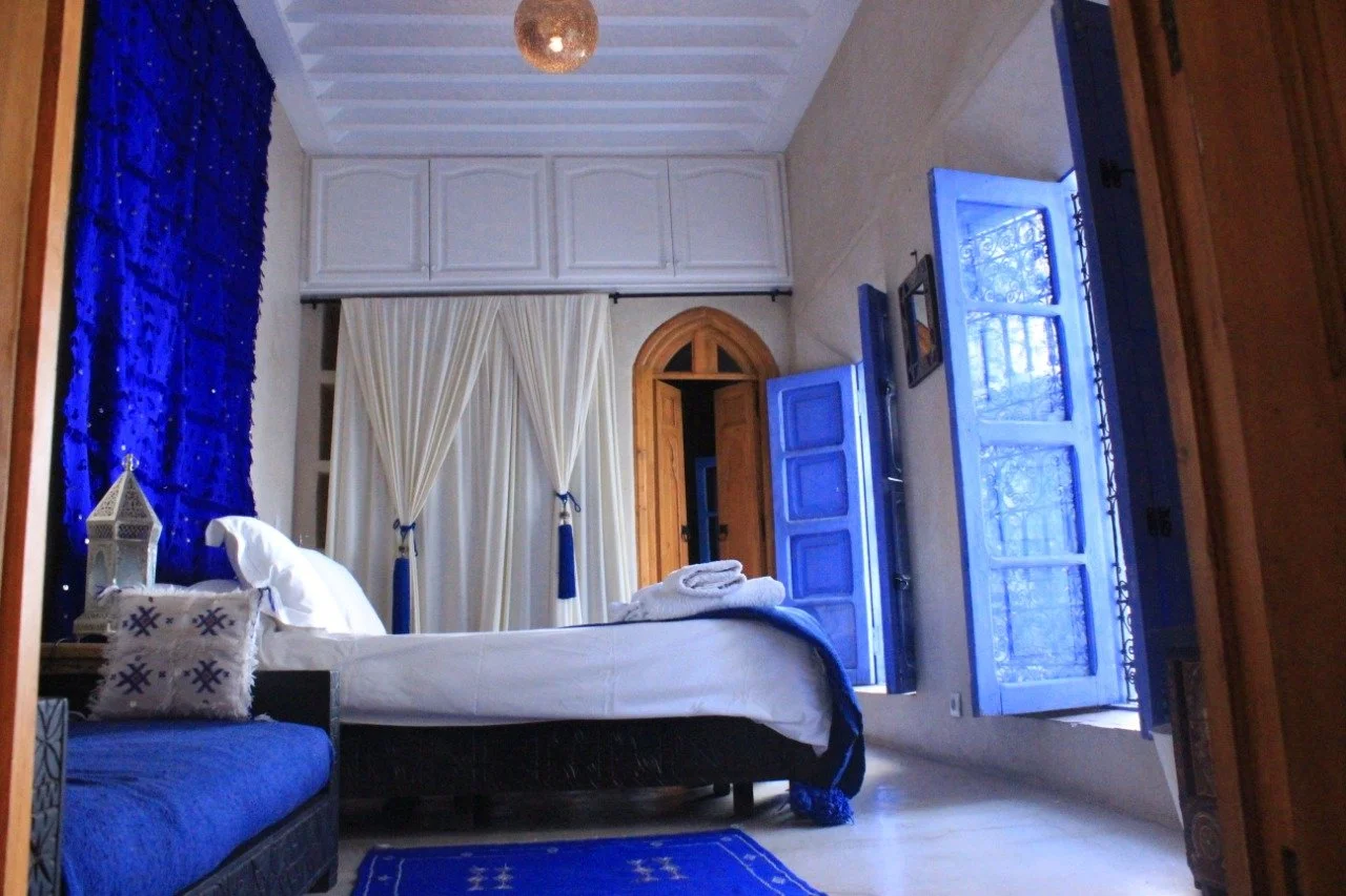A bedroom with a bed, blue and white curtains, a blue couch, and open blue windows with a view of trees outside.