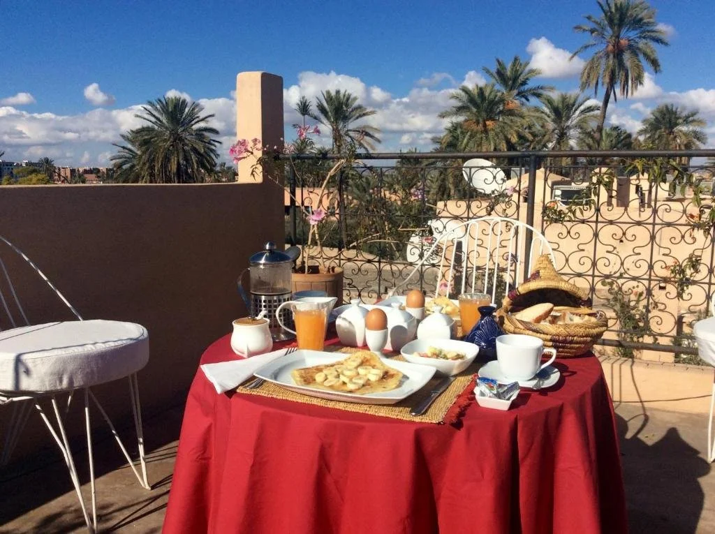 Breakfast table with a red tablecloth featuring pancakes, eggs, toast, orange juice, coffee, and sugar, set on a rooftop with palm trees and a blue sky in the background.