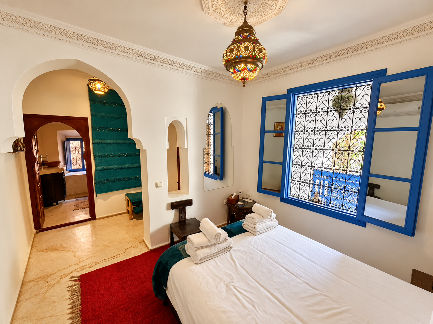 A bedroom with a white bed, folded towels, and blue framed windows with iron bars, in a room with Moroccan-style lantern overhead and arched doorways.