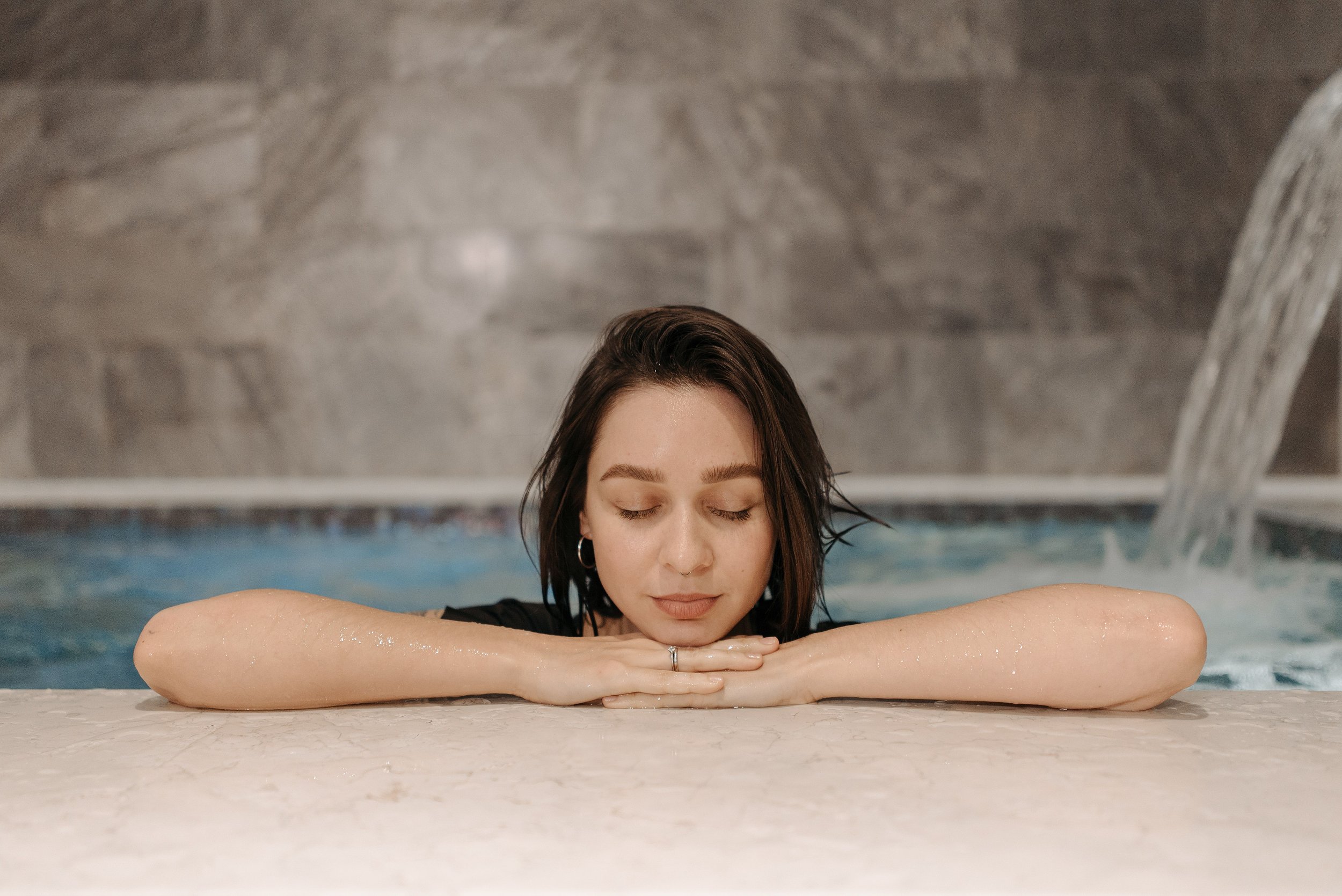 A woman with closed eyes and wet dark hair resting her arms and head on the edge of a swimming pool with her hands clasped.