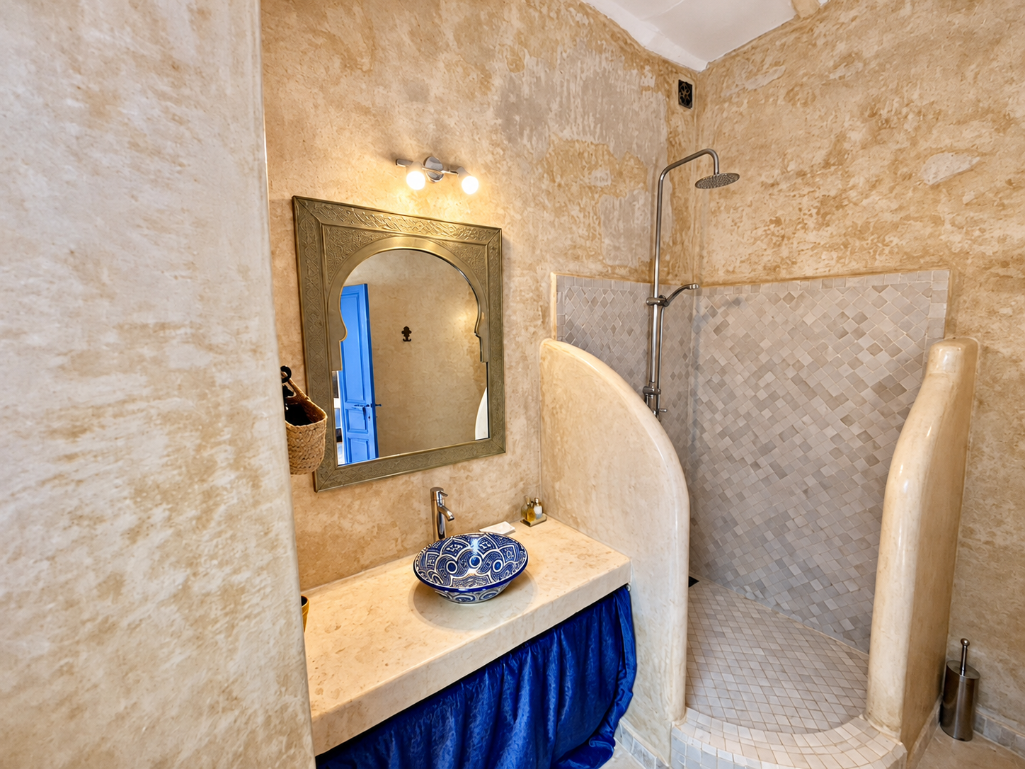 Cozy bathroom with stone walls, a shower area with light-colored tile, a decorative mirror, and a vessel sink on a marble countertop with a blue curtain beneath.
