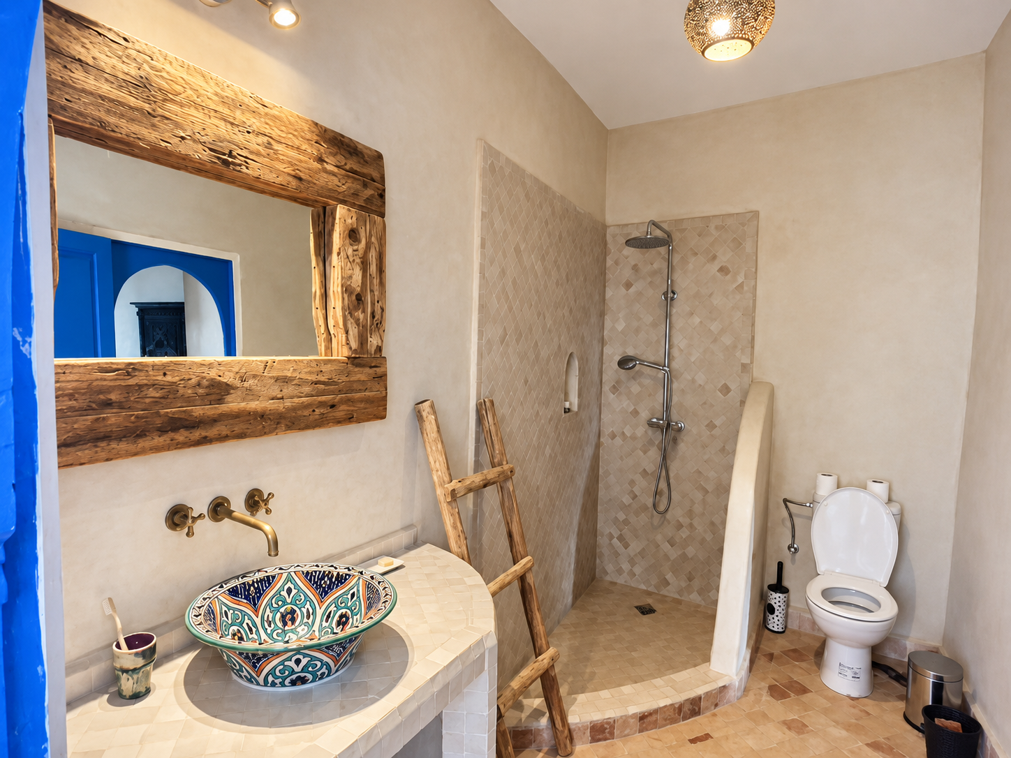 A bathroom with a rustic wooden-framed mirror above a decorative tile vessel sink, a brass faucet, a wooden ladder leaning against the wall, a walk-in shower with beige tile walls, a handheld showerhead, and a small niche, a white toilet with two roll holders, toilet paper, a black toilet brush, and a small trash bin.
