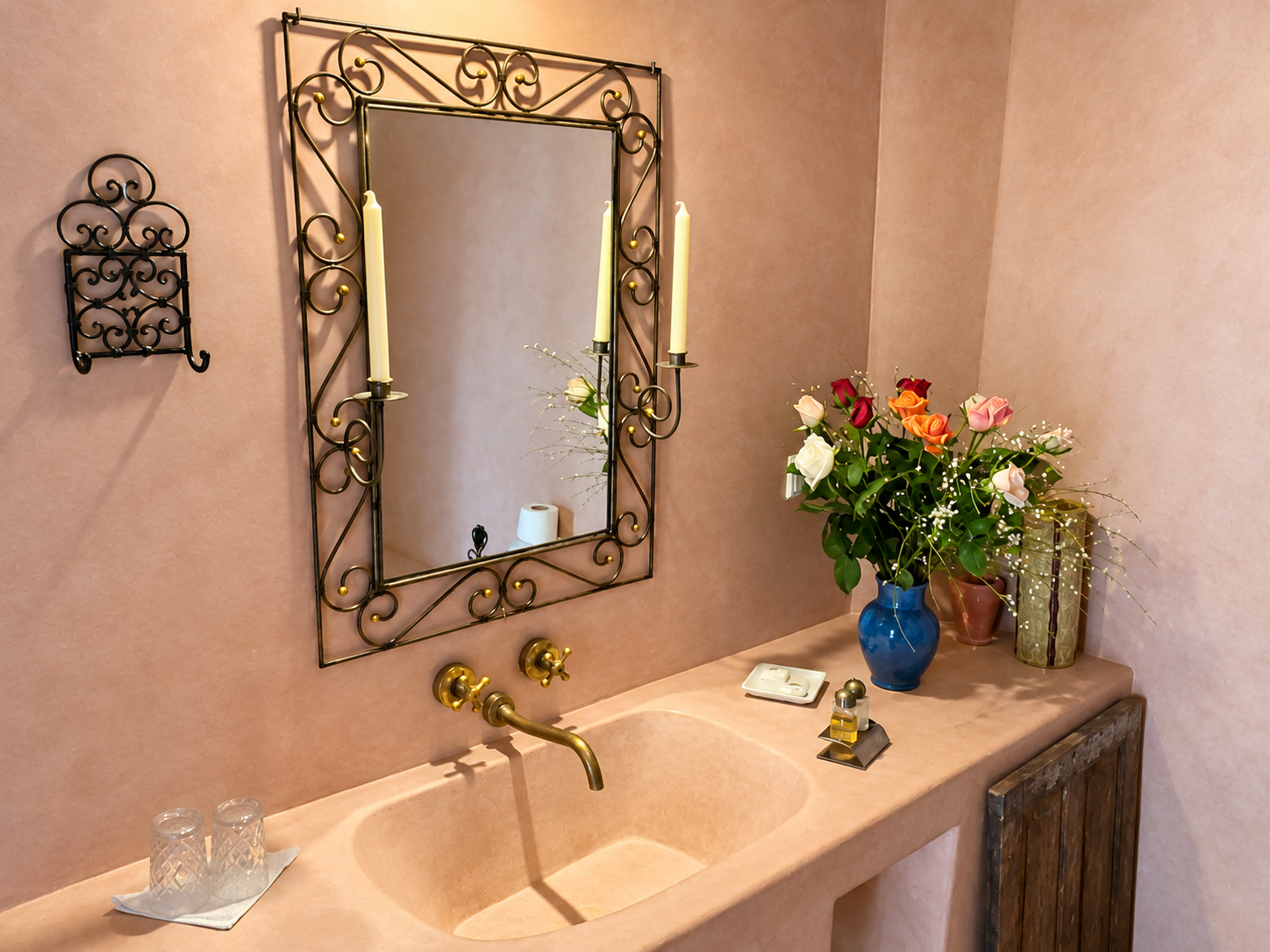 A bathroom vanity with a pink countertop, gold faucet, and a large mirror with an ornate black and gold metal frame. Next to the mirror is a wall-mounted decorative black metal rack. On the countertop are two glasses on a tray, a white soap dish, a small bottle of perfume, and a large blue vase filled with pink, white, and orange roses along with greenery.