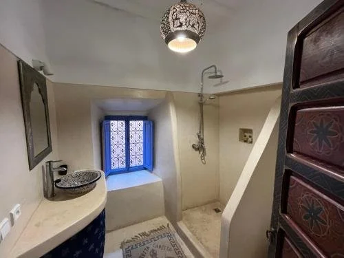 Bathroom with a small blue window, shower area, decorated ceiling light, and a curved countertop with a sink, storage cabinet, and decorative items.