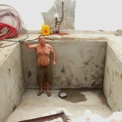 A shirtless man standing inside a large empty concrete rectangular pool under construction, with construction tools and equipment around.