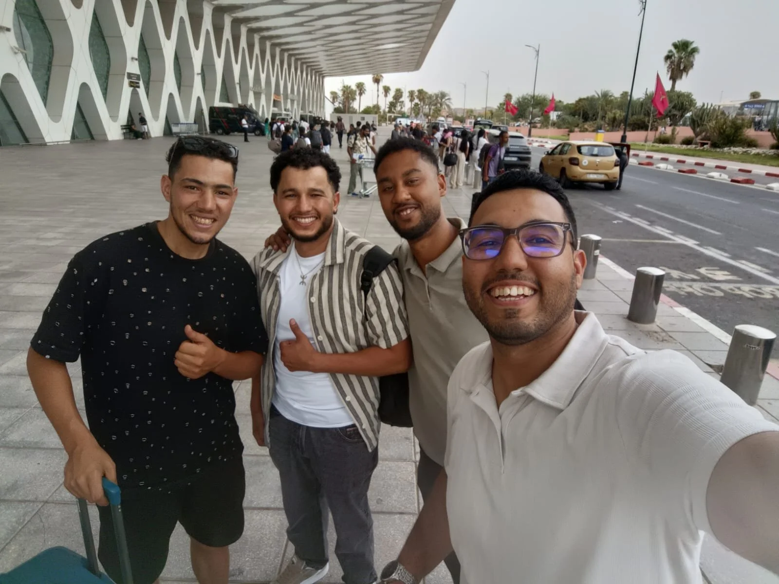 Four smiling men taking a selfie outside an airport building with people, cars, and palm trees in the background.