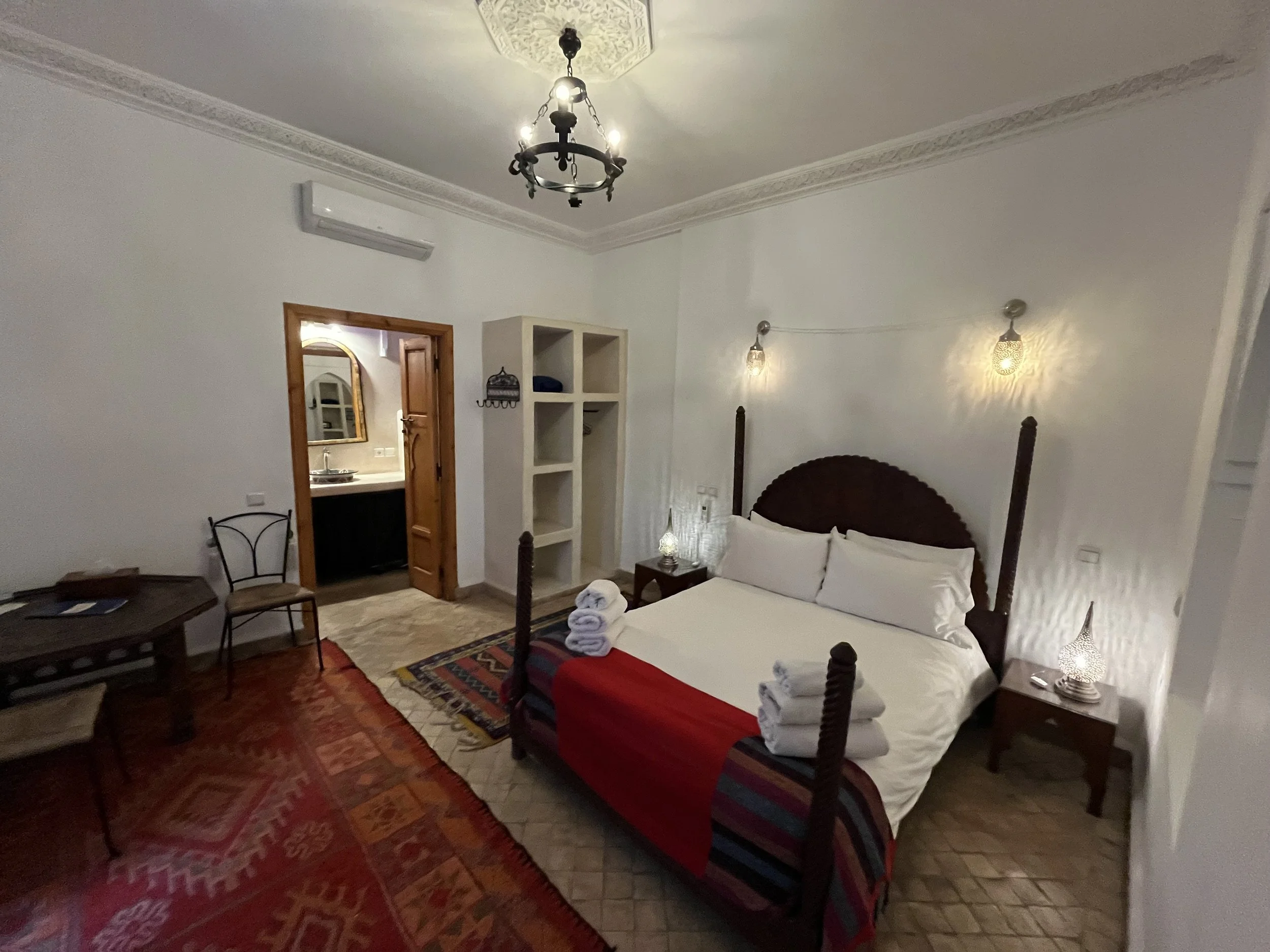 A bedroom with a wooden four-poster bed covered with white bedding and folded towels. There is a table with a lamp, a shelf unit, a wooden door, and a small sitting area with chairs and a table. The room has white walls, decorative crown molding, and hanging wall lights. The ceiling features a black chandelier and an ornate ceiling medallion.