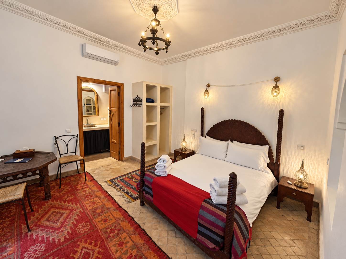 A hotel room with a queen-sized bed, two bedside tables with decorative lamps, a small shelf, and a wooden table with chairs. The room features white walls, ornate ceiling molding, and a chandelier. Towels are folded on the bed, and a red and multicolored rug covers part of the tiled floor. An open door reveals a bathroom with a sink and mirror.