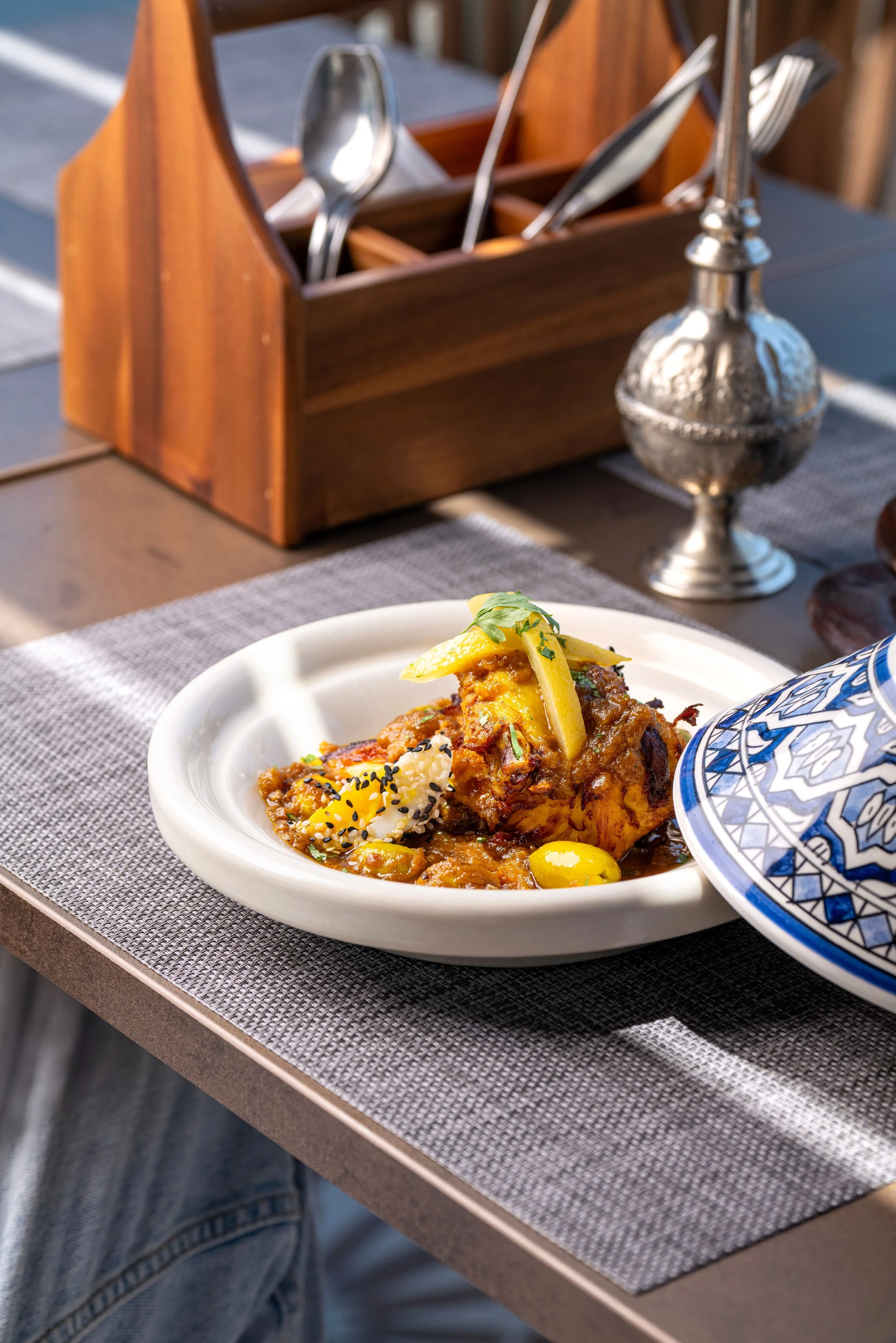 A white bowl of stew garnished with a lemon wedge, herbs, and black sesame seeds, next to a blue and white patterned ceramic bowl, on a gray placemat at an outdoor table.