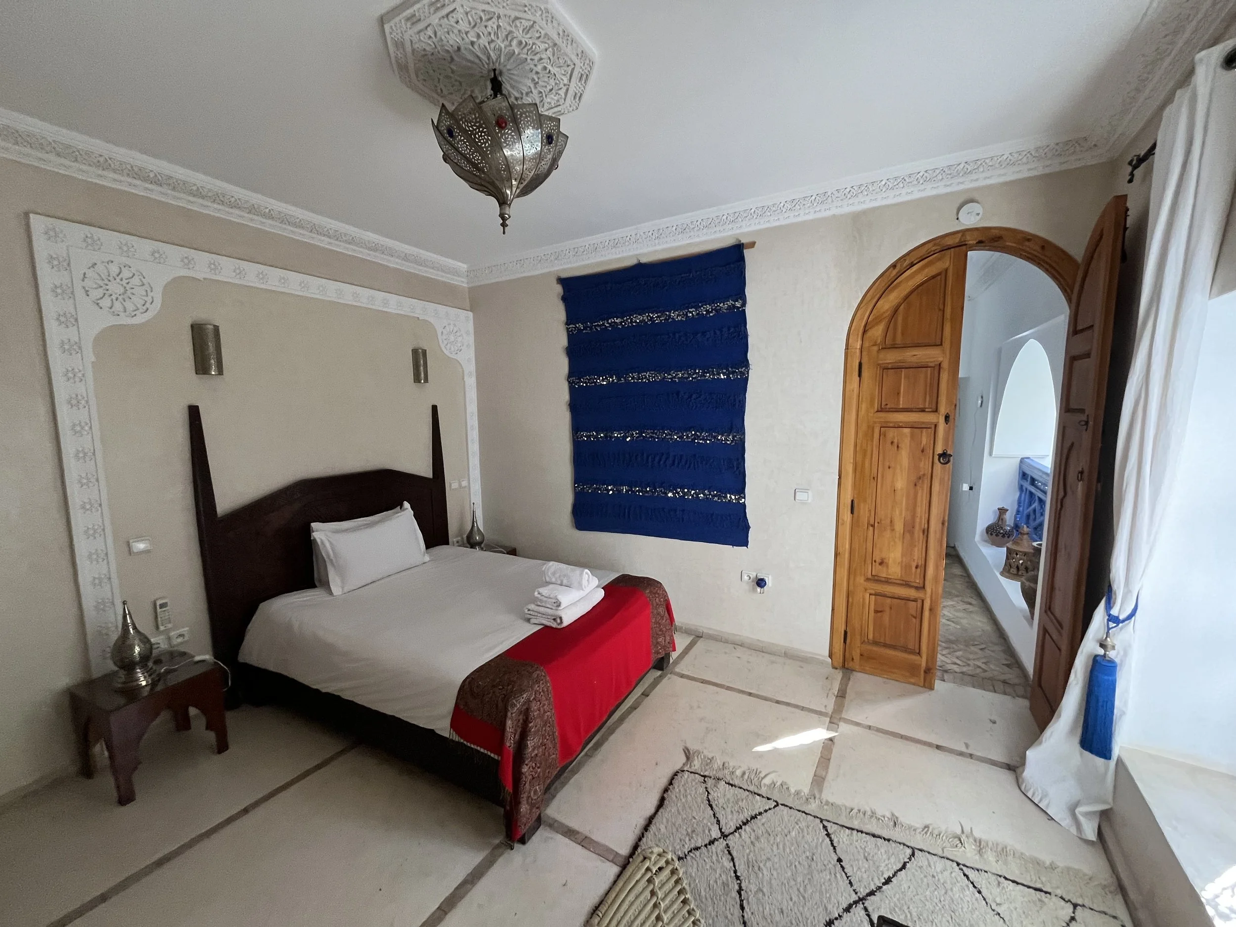 A bedroom with a bed, red and gold bedspread, towels, nightstands, silver lamps, blue tapestry, wooden door, and white curtains.
