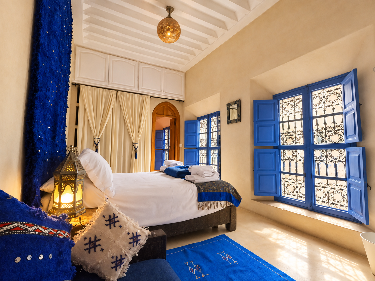 A bedroom with a white bed, blue accents, and Moroccan-inspired decor. Features include blue shutters on windows, an ornate lantern on a bedside table, and beige curtains near an arched doorway.