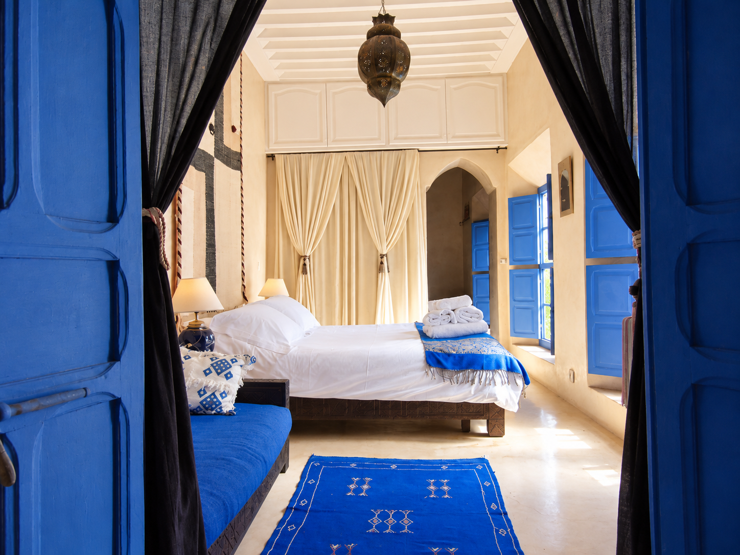 Interior view of a bedroom with blue doors and window shutters, a bed with white linens and rolled towels, beige curtains, a blue rug, and traditional lantern ceiling light