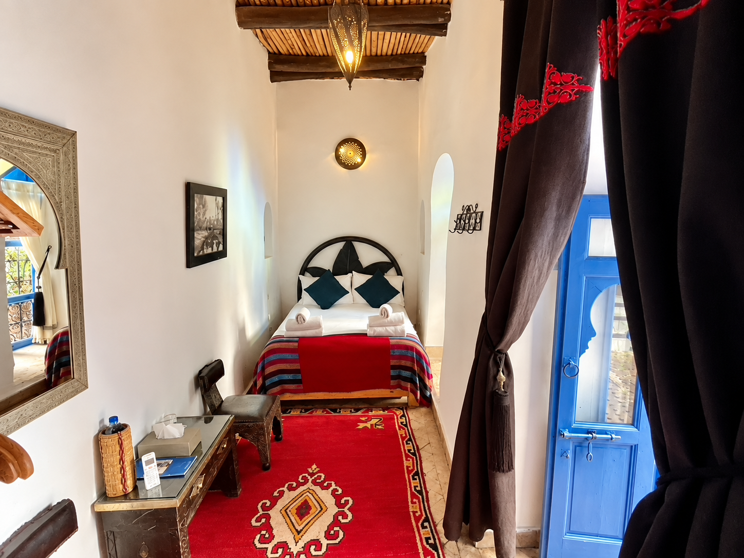 A cozy bedroom with a double bed decorated with dark headboard, white bedding, three teal pillows, and two rolled towels. The bed has a colorful striped blanket. The room features white walls, a wooden ceiling with exposed beams, and a red patterned rug. To the right, there is a blue door with a curtain partially open, revealing natural light. On the left, a small table with a mirror, some items, and a chair are visible.