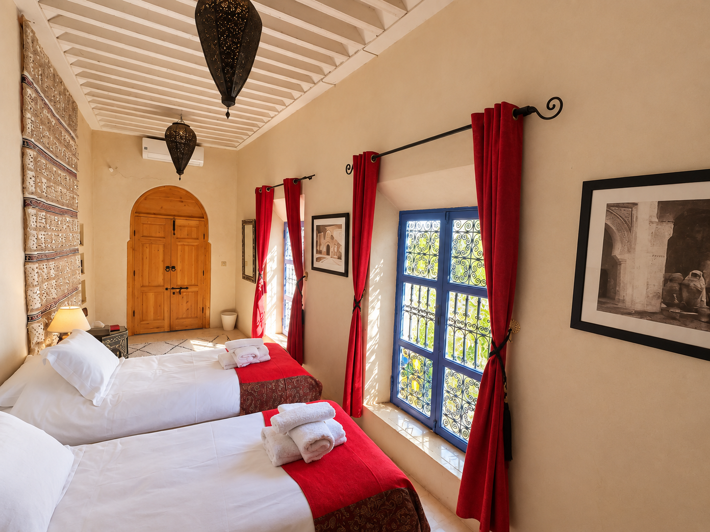 A cozy bedroom with two white beds, each with neatly folded towels on top. The room has a rustic style with Arabic-inspired decor, including a tapestry on the wall, red curtains, and framed black-and-white pictures. There are two blue glass-hinged windows with intricate black wrought iron grilles, allowing sunlight to brighten the room. The ceiling has exposed white wooden beams and hanging Moroccan-style lanterns, with a wooden door at the back of the room.