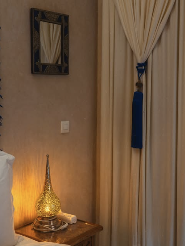 A bedside lamp shaped like a teardrop with a warm glow, placed on a wooden nightstand next to a bed with a white pillow. Decorative curtains are partially closed, tied back with a blue ribbon, covering a window. A mirror with a decorative frame hangs on the wall above the nightstand.
