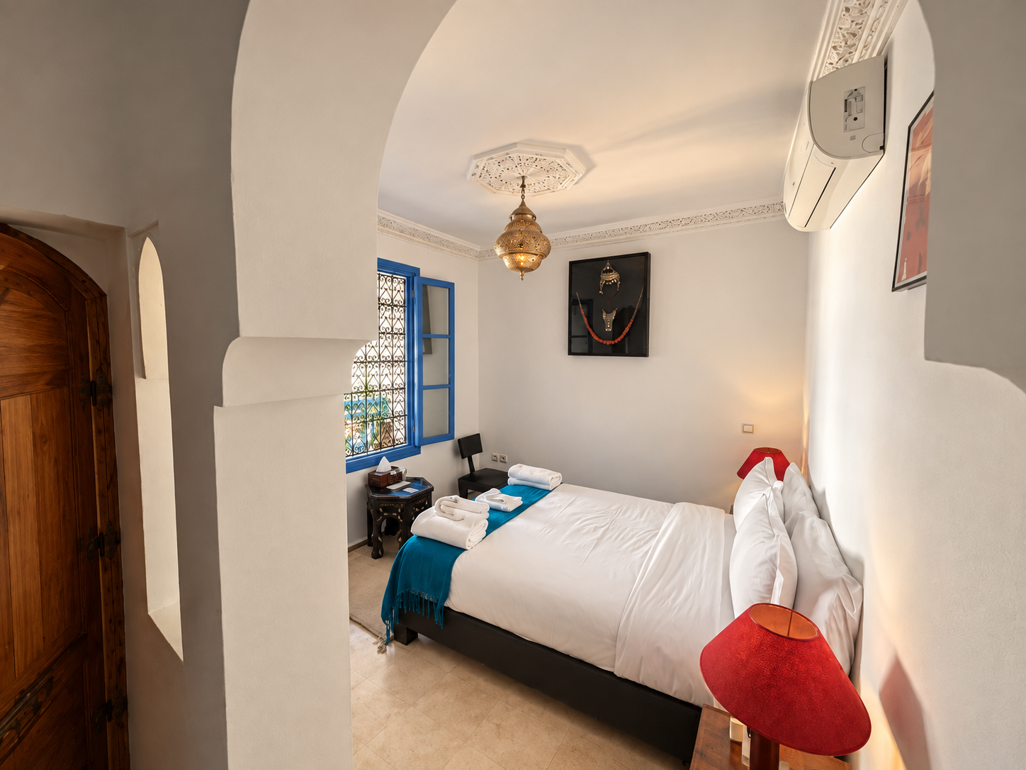 A bedroom with white walls, a large bed with white linens, a red bedside lamp, and a blue throw blanket. There are two towels on the bed. A window with blue framing and ornate ironwork is on the left, with a view outside. A decorative hanging lamp is on the ceiling, and a framed Moroccan-style wall art hangs above the bed. An air conditioning unit is on the right wall.
