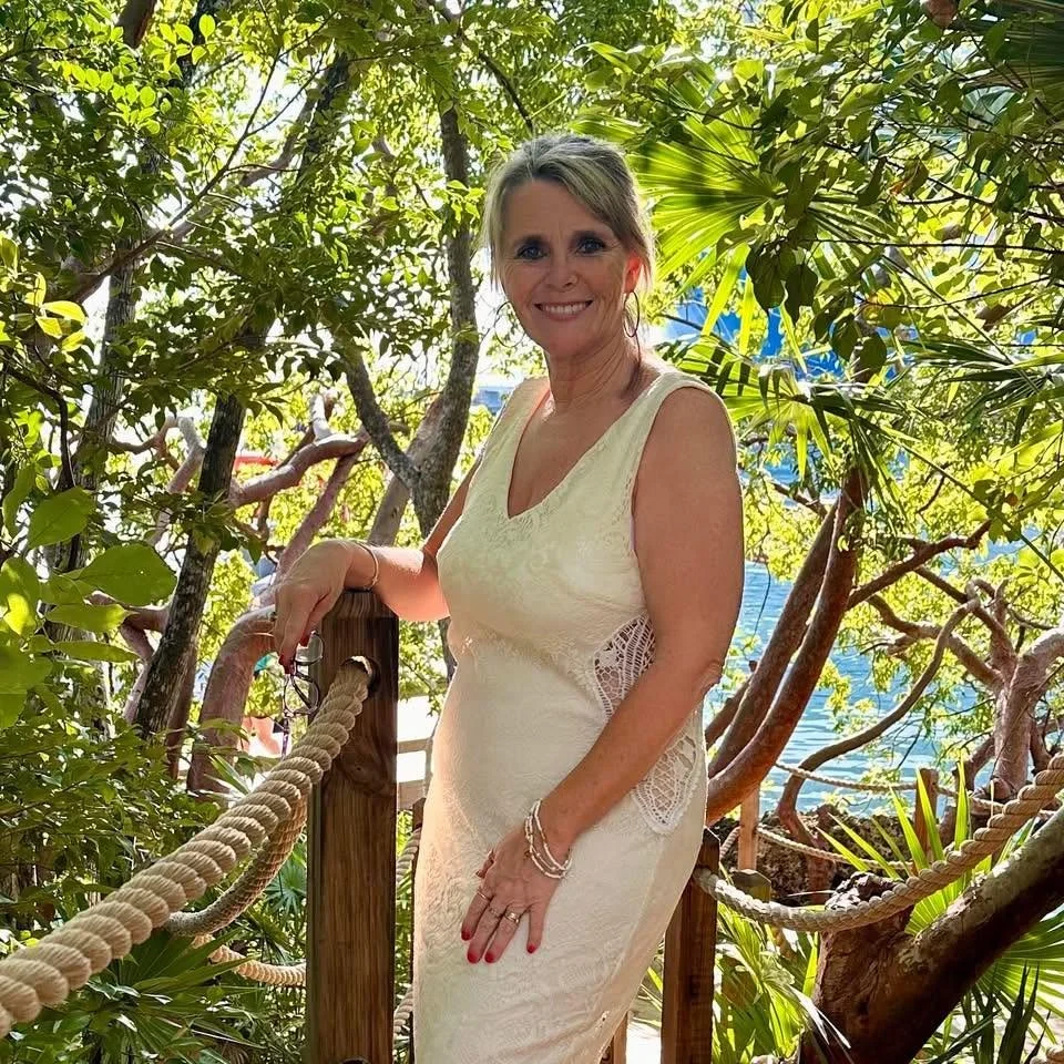 A woman in a cream-colored dress standing on a wooden deck with a rope railing surrounded by lush green trees and shrubs, with water visible in the background.