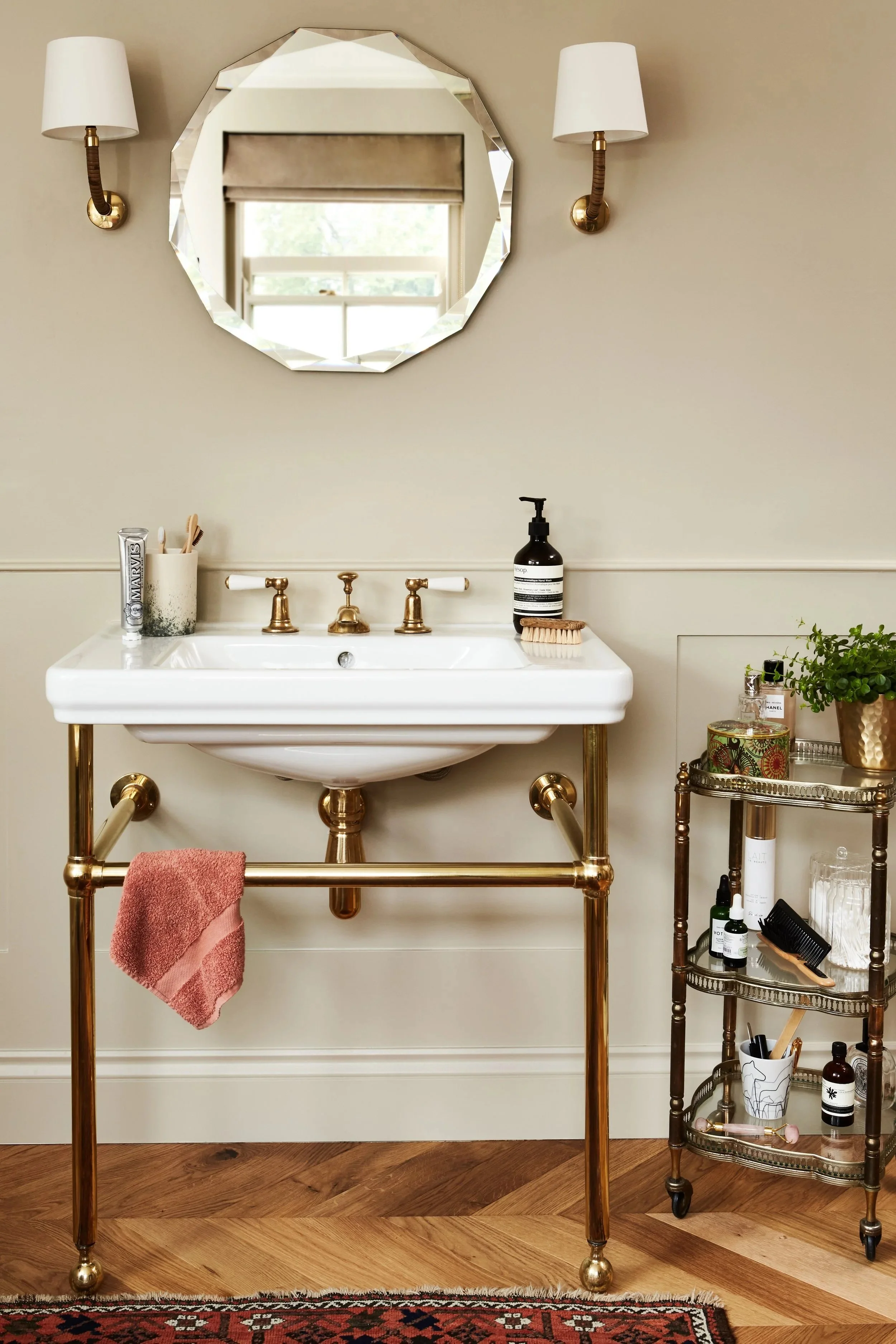 Elegant bathroom vanity with white sink, brass fixtures, faceted mirror, wall sconces, toiletries, and a pink hand towel