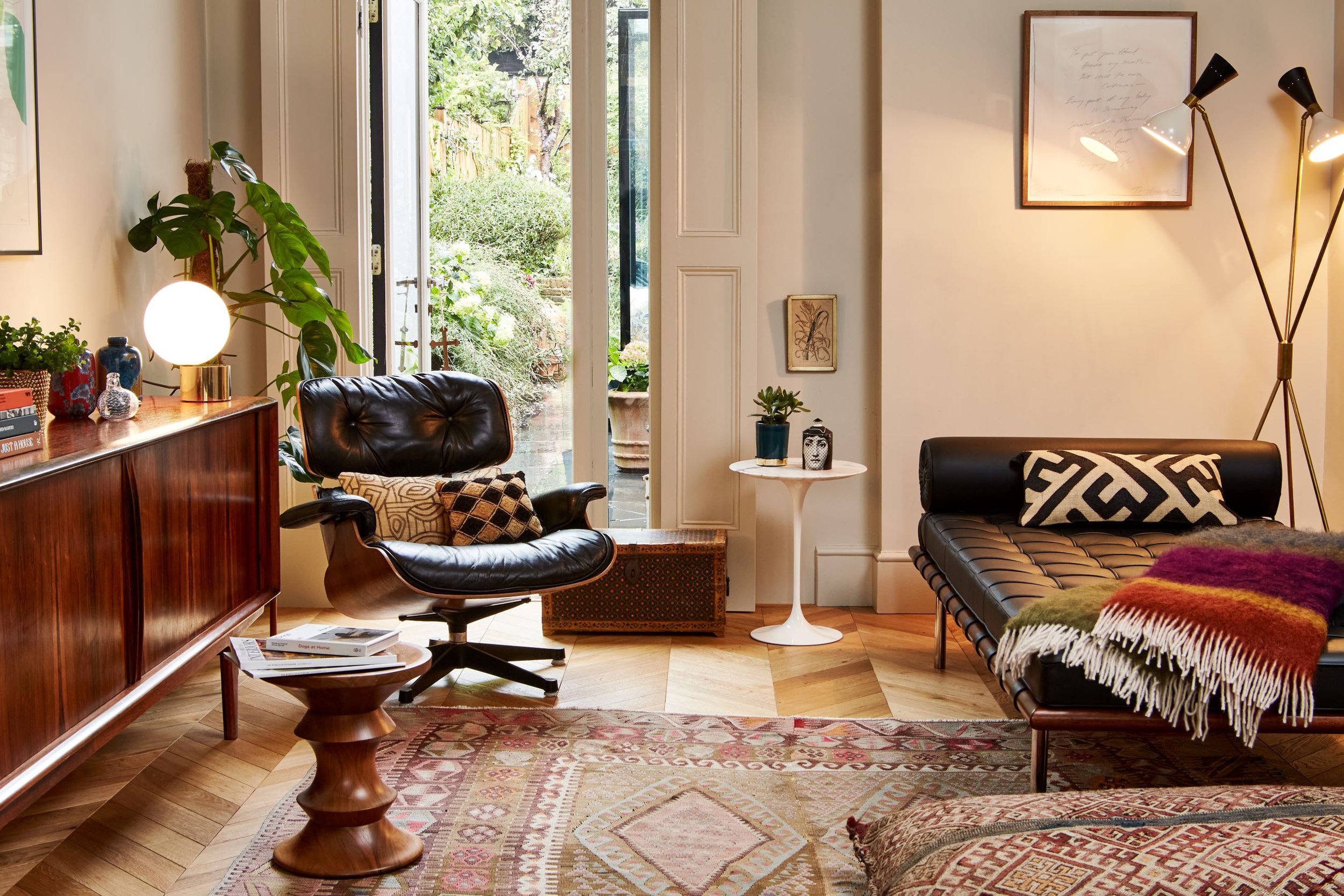 Living room with wooden furniture, black leather armchair, black leather sofa, small white side table with potted plant and decorative items, open door leading to a garden, patterned rugs, framed artwork, inside standing lamp with multiple adjustable