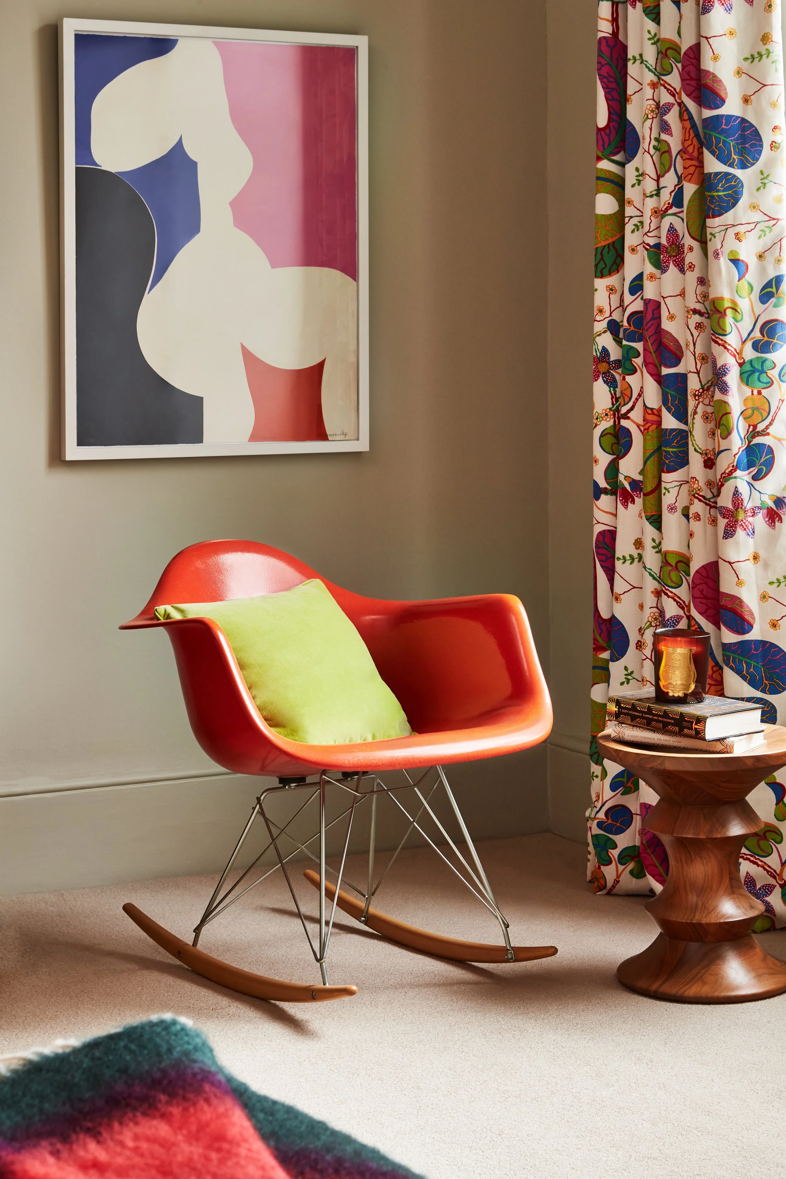Colorful mid-century modern reading nook with a orange rocking chair, green cushion, abstract framed artwork, Josef Frank patterned curtains, books, and eames wooden side table.