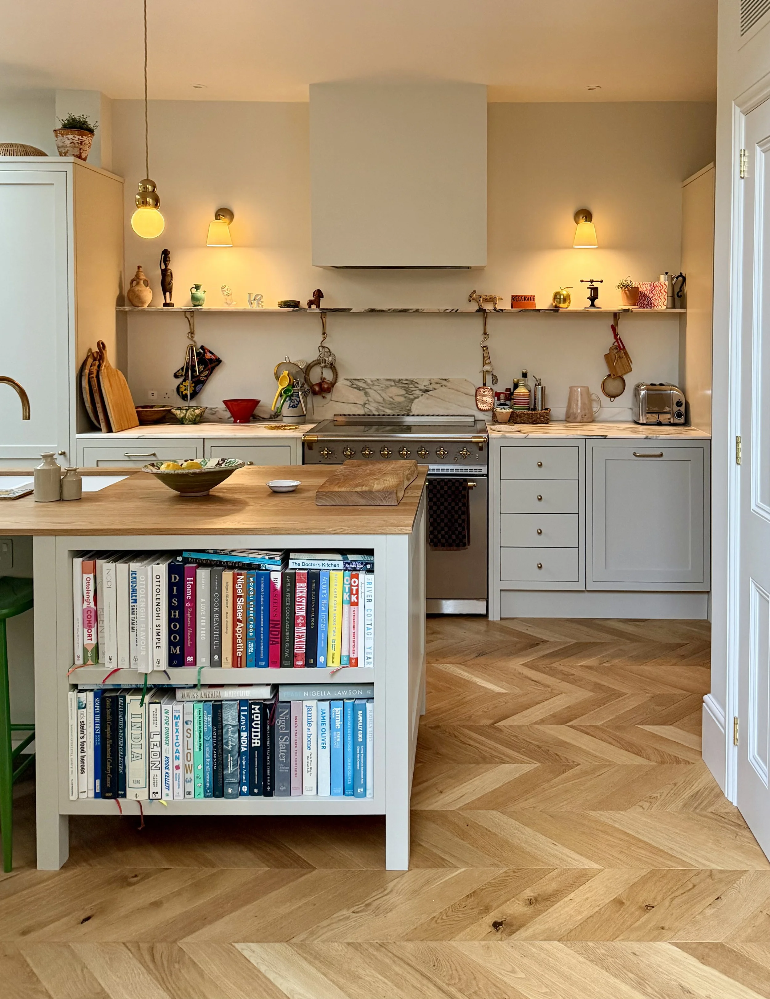 A kitchen with a white island that has a bookshelf filled with books on one side. The kitchen has white cabinets, a marble backsplash, and wooden flooring. The shelves above the stove hold decorative items, plants, and a small sign. The countertop ha