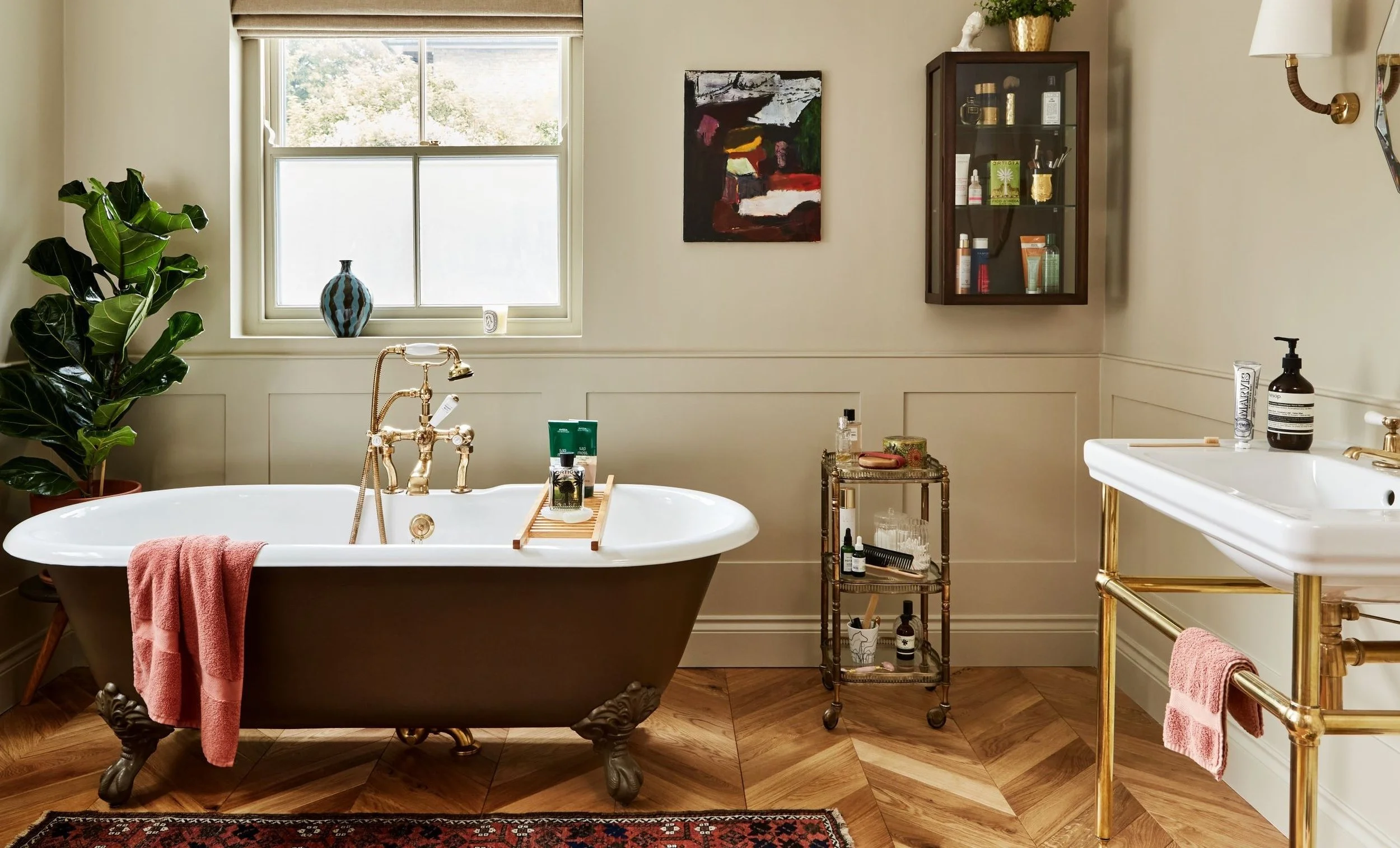 A bathroom with a large window, a vintage clawfoot bathtub with a pink towel, green plants, a small side table with toiletries, and a white sink with gold legs. There is an abstract painting and a glass cabinet on the wall.