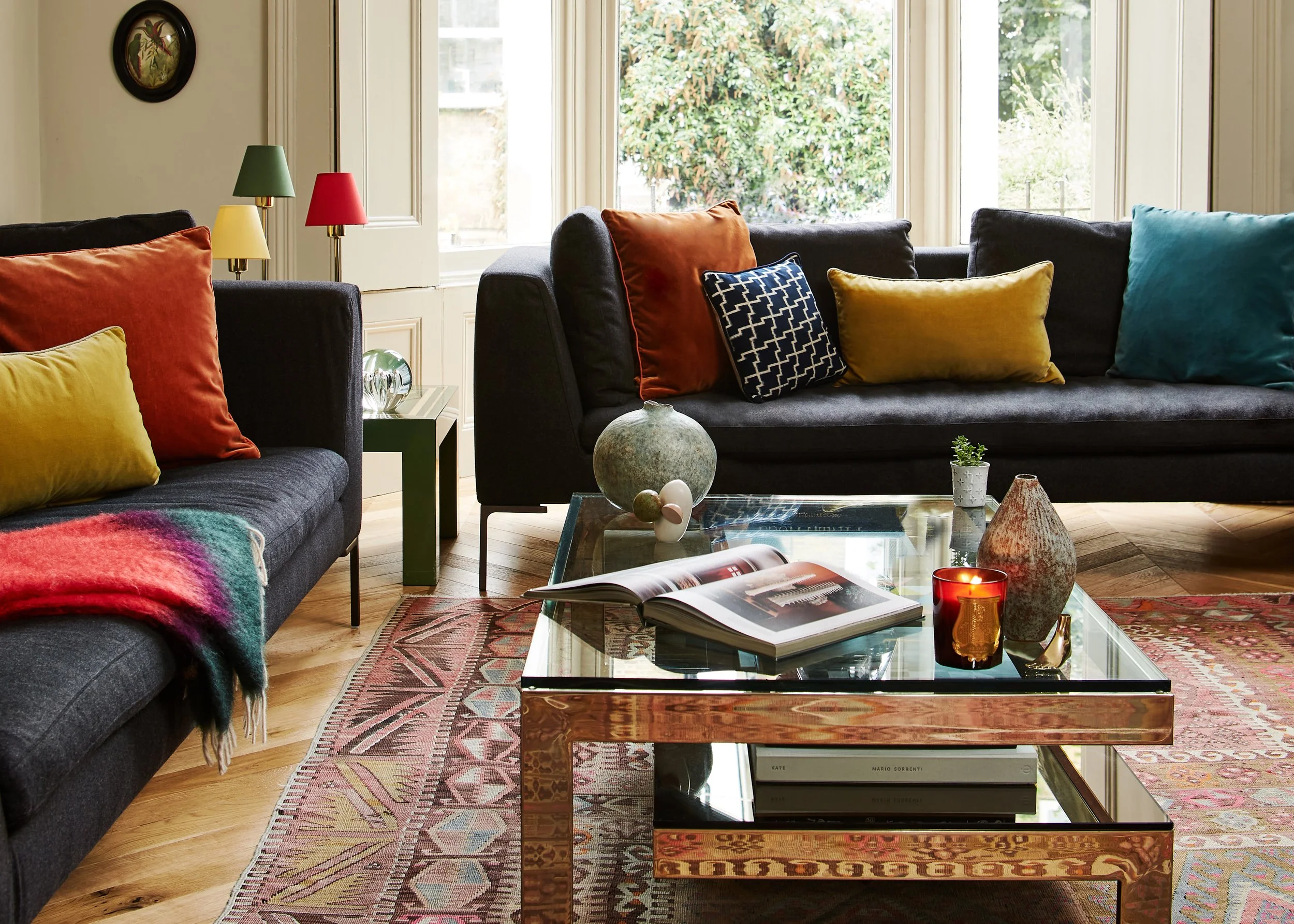 Living room with dark gray sofas, colorful pillows, a glass coffee table with books and decorative objects, a patterned rug, and large windows with greenery outside.