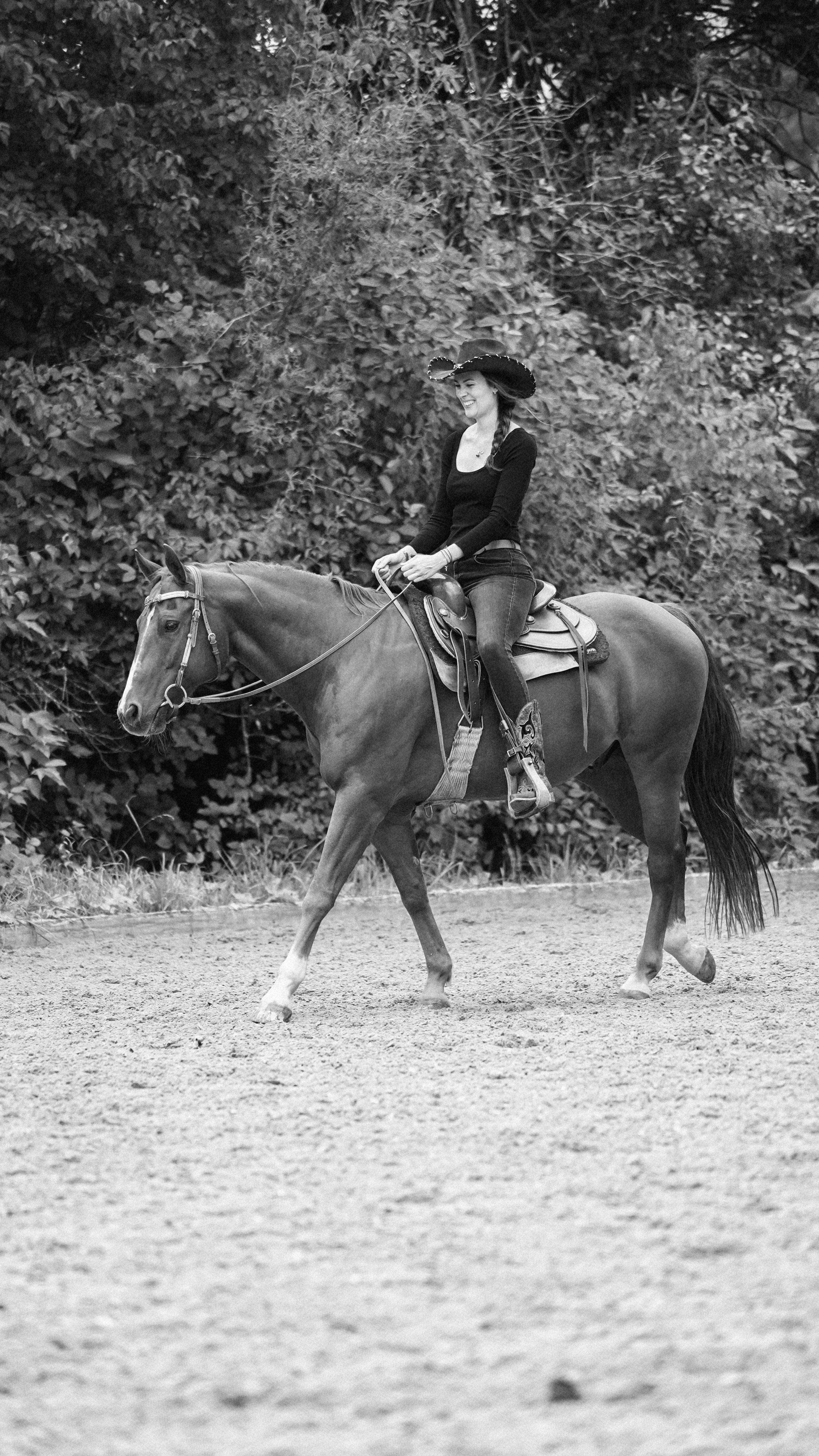 Die Schwarz-weiß Fotografie zeigt eine Frau mit einem Cowboyhut und Westernstiefeln, die auf einem Pferd auf einem Sandplatz, am Rande eines Waldes reitet.