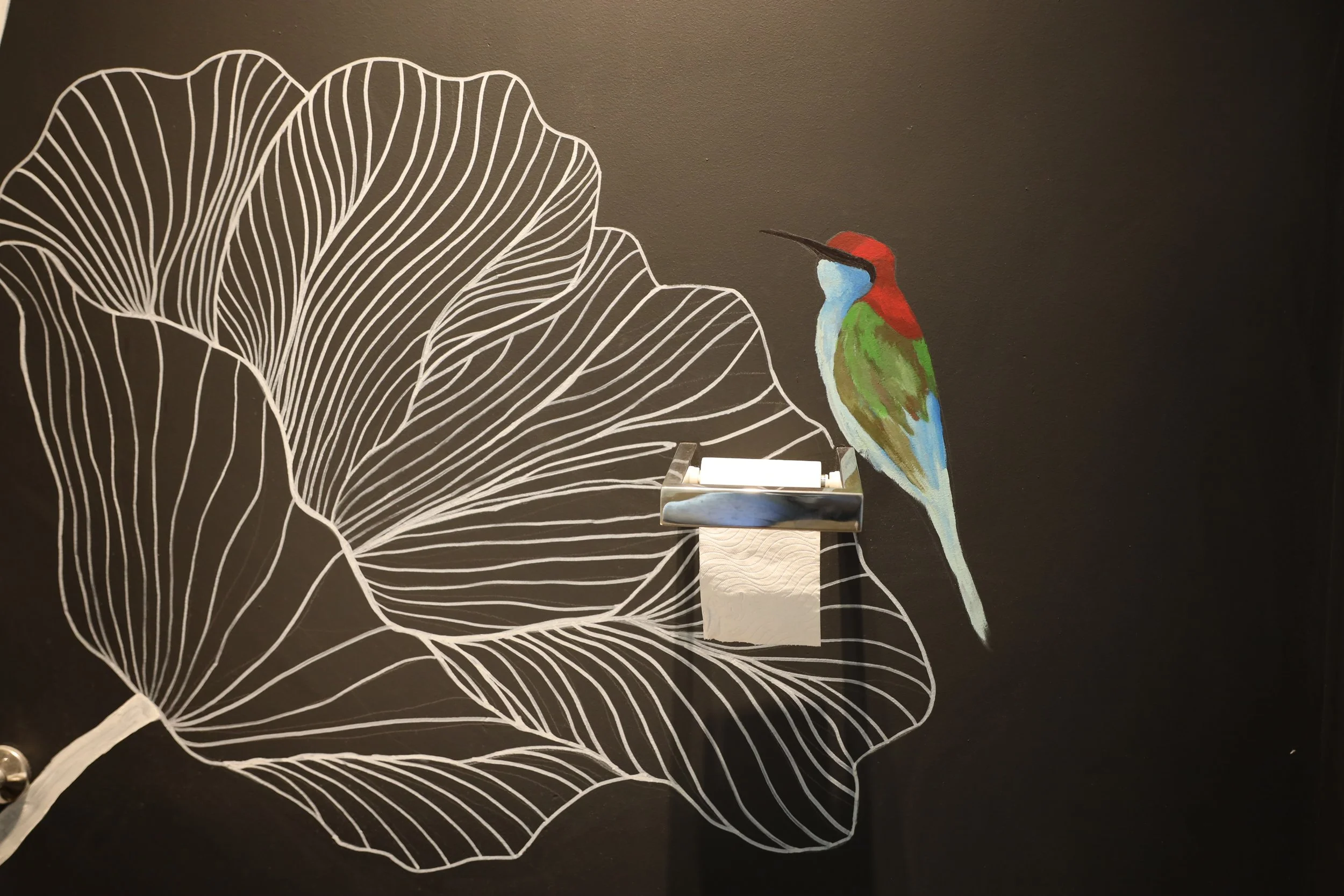 A mural of a colorful bird, likely a kingfisher, perched on a branch with a background of large, white outline leaves on a dark wall.