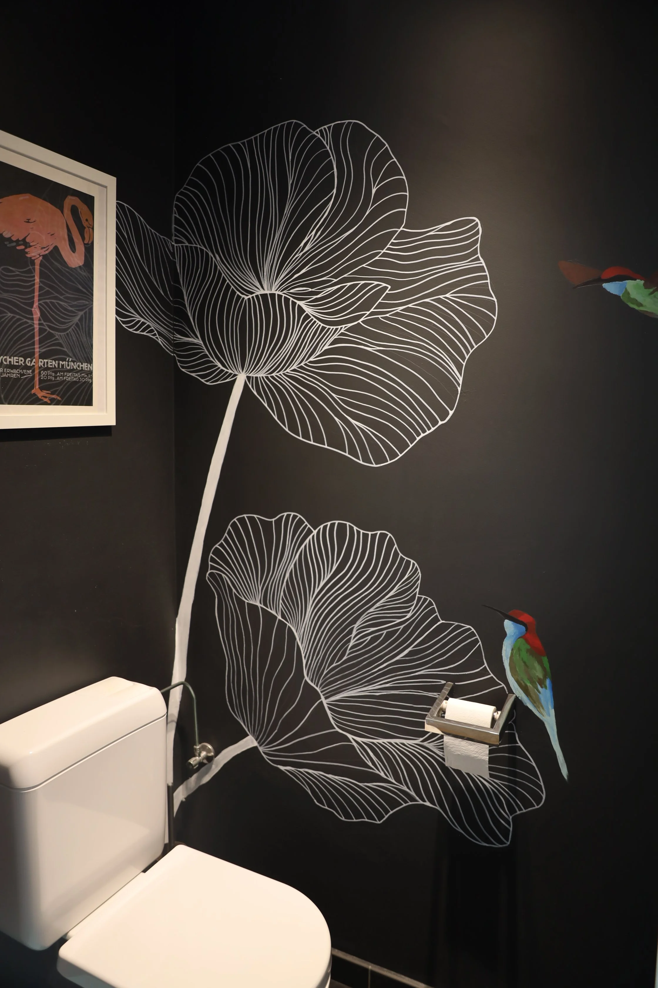 Black bathroom wall with large white line art floral design and a colorful hummingbird stencil. A framed flamingo poster on the wall. A white toilet and toilet paper holder are present.