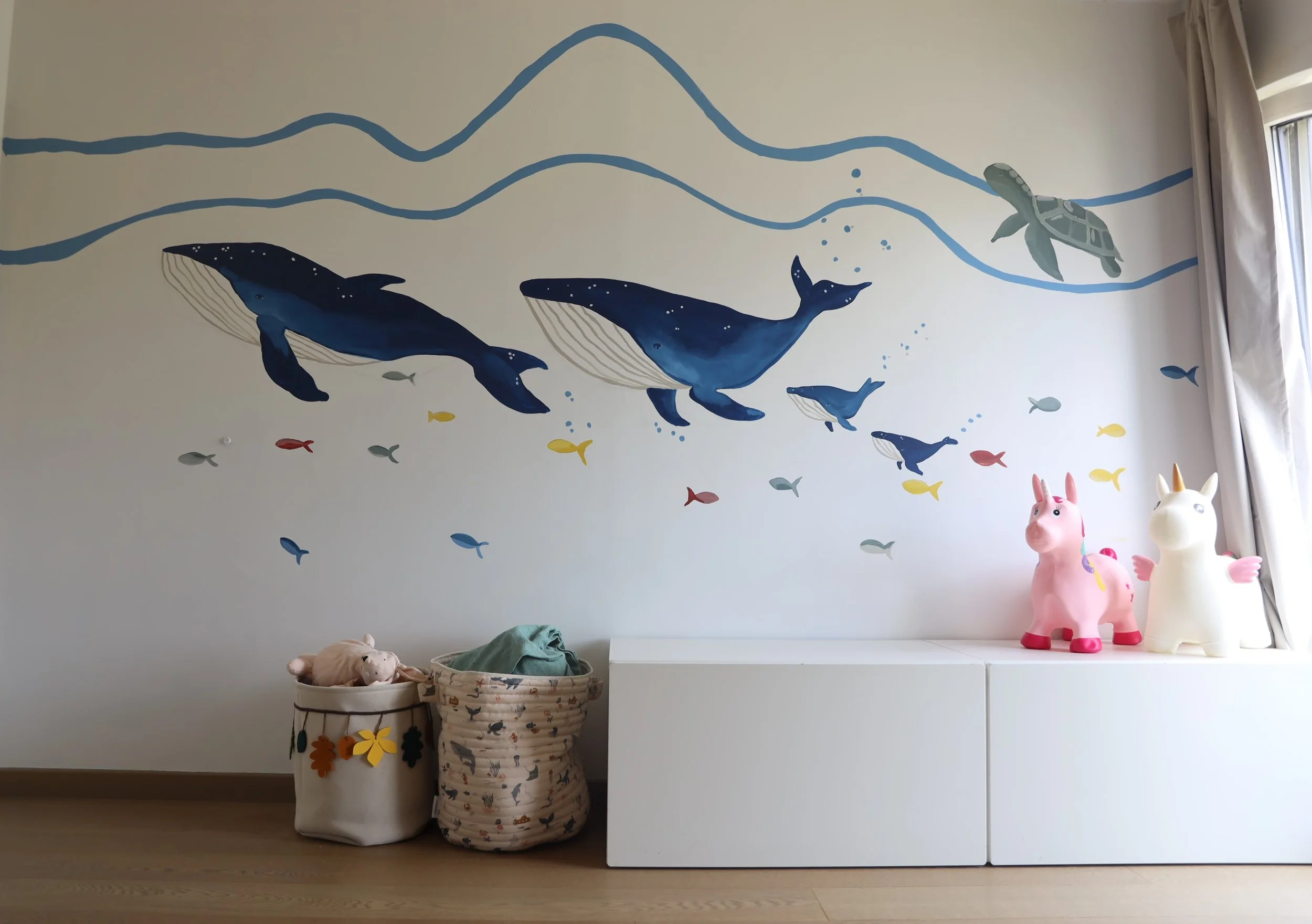 A wall mural of blue whales swimming with a sea turtle and various small fish in a children's room. Below the mural, there are two fabric storage baskets filled with plush toys, and on a white cabinet, there are two cartoon pig and unicorn toys.