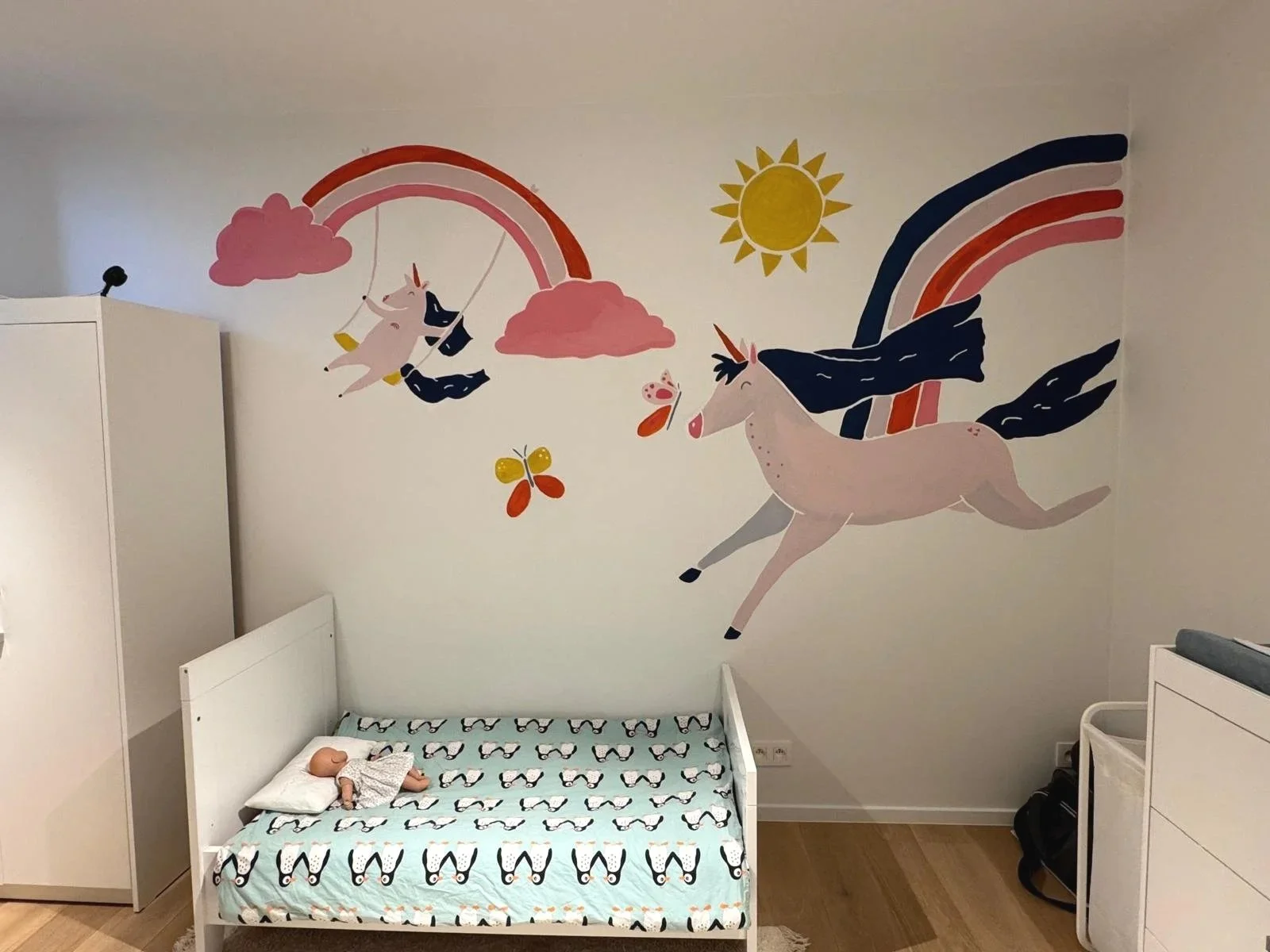A children's bedroom with a wall mural featuring a unicorn, rainbows, clouds, a sun, a butterfly, and a flying unicorn with a rainbow.