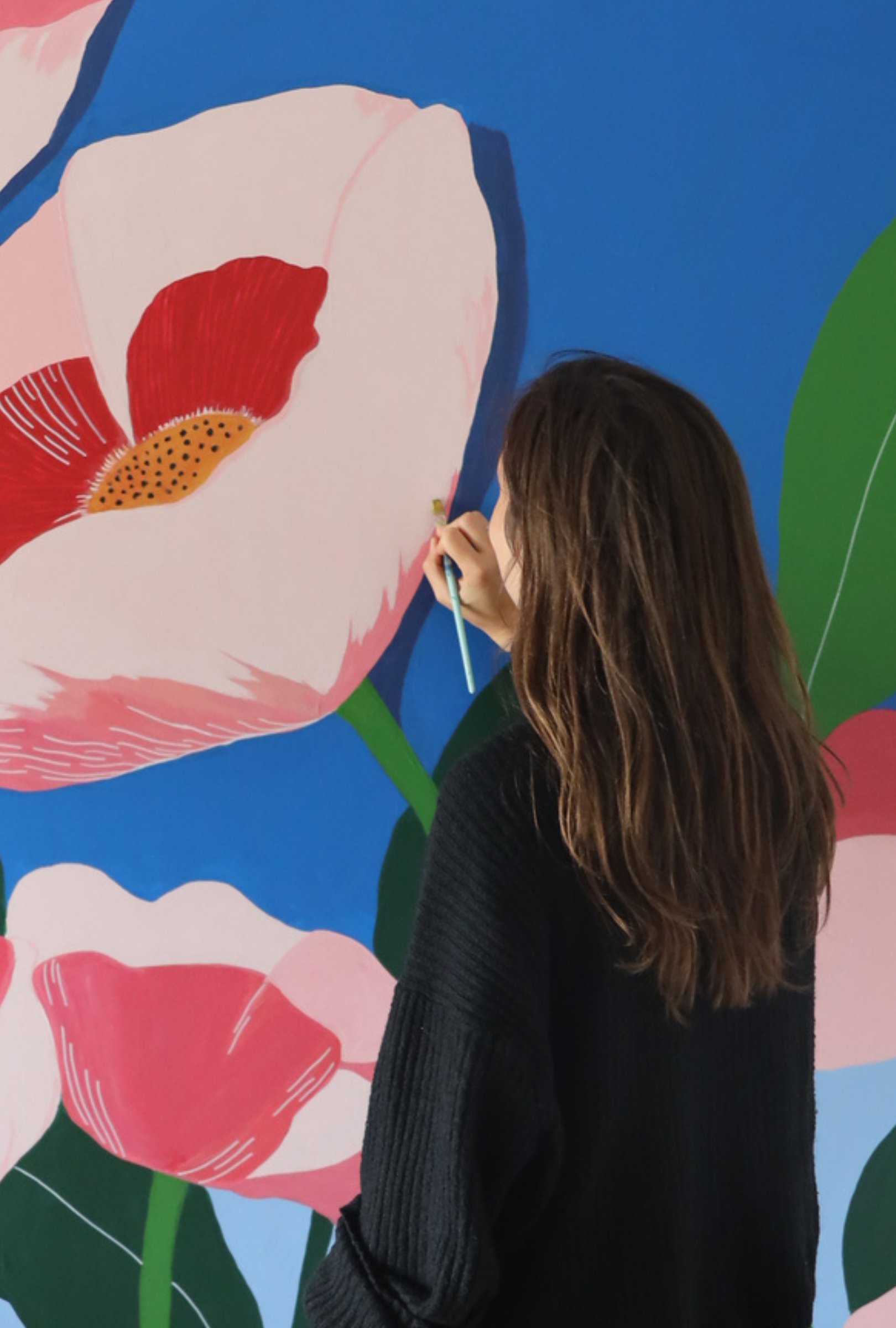 A woman with long brown hair painting a large floral mural with pink and white flowers and green stems on a blue background.