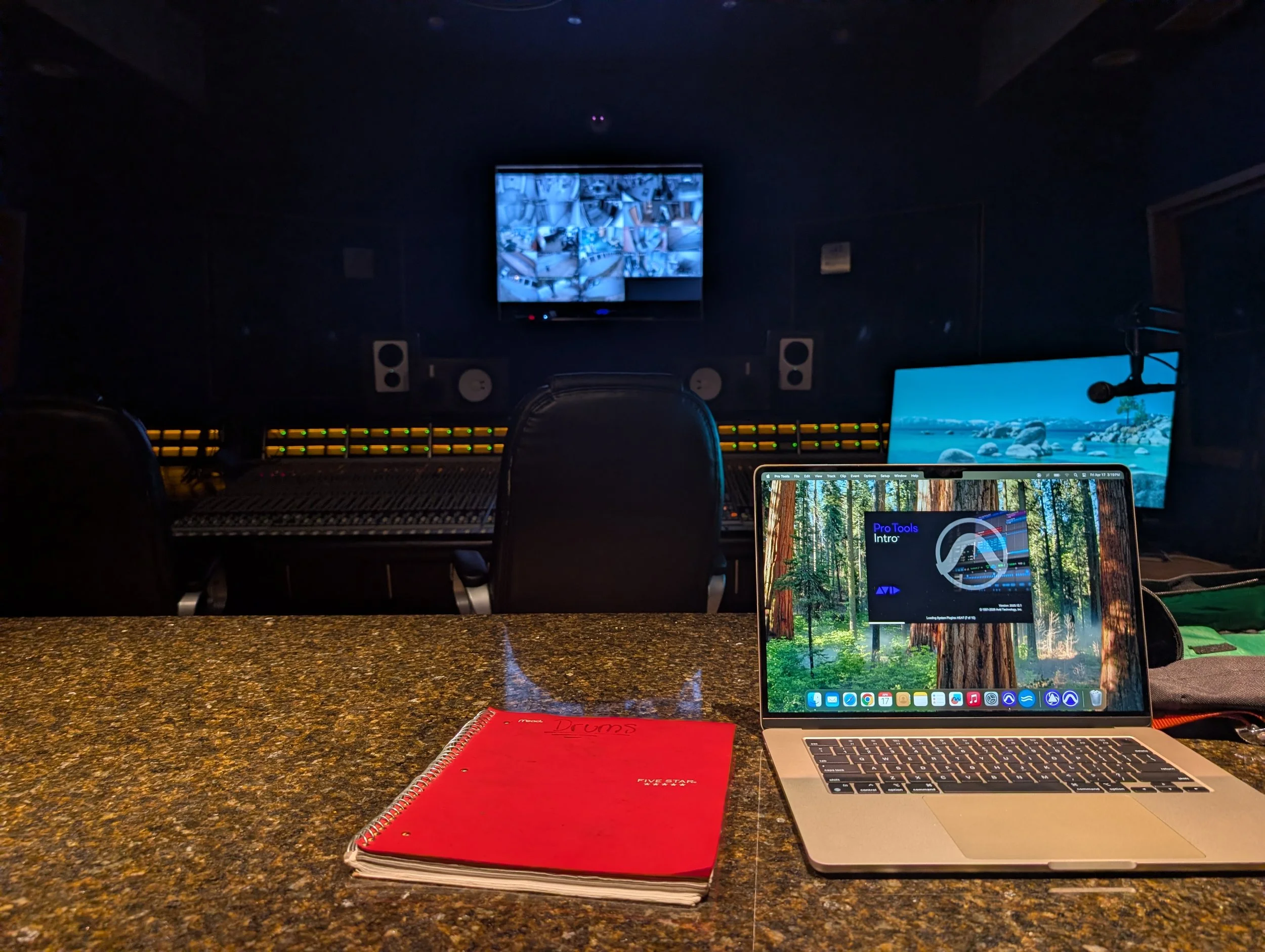 Music recording and production studio with mixing console, computer, and monitors in a dark room, viewed from a granite countertop.